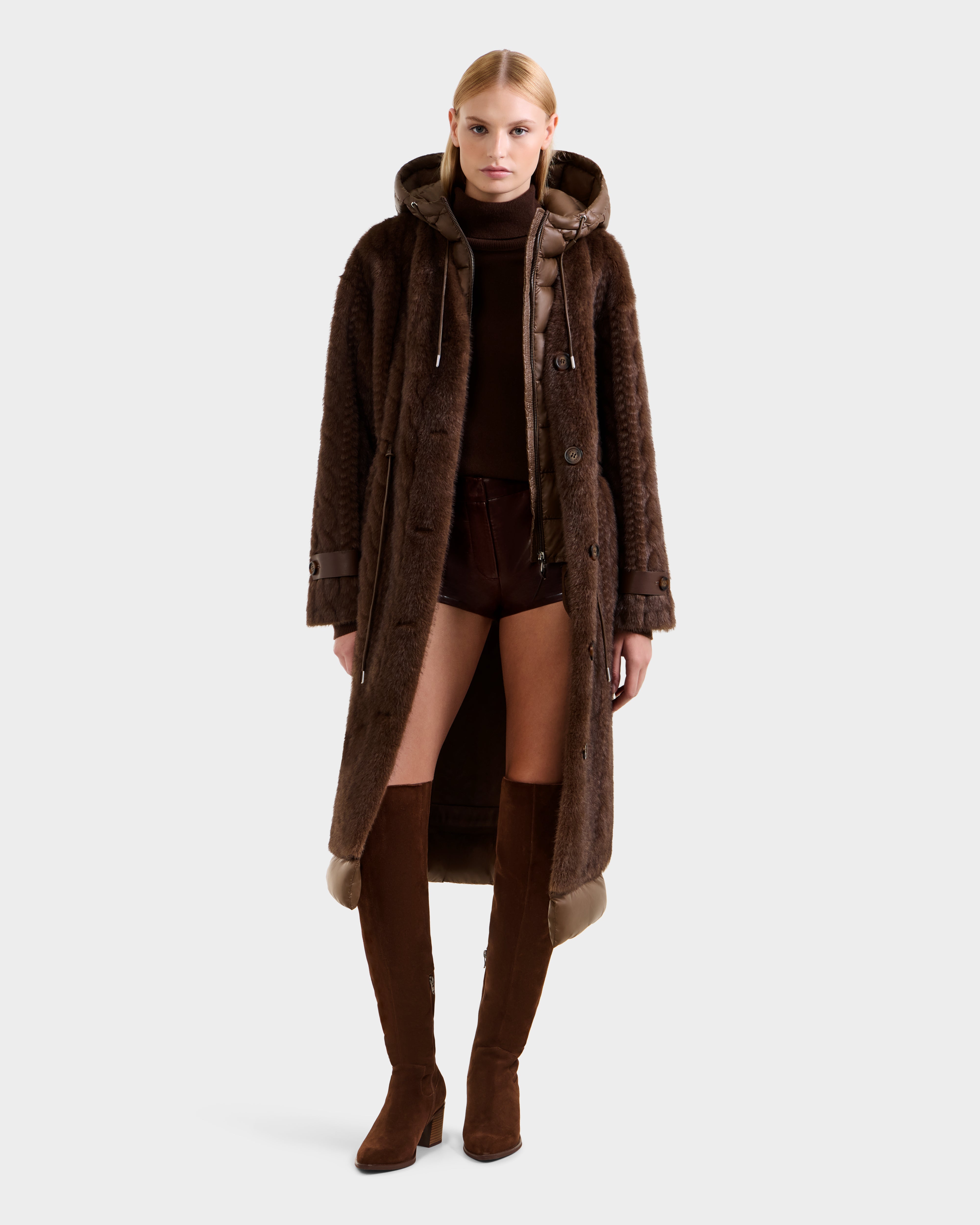 A woman faces forward, wearing the Rudsak ORIANA - 8125545 Mocha faux fur hooded coat with a dark brown turtleneck, brown shorts, and knee-high brown suede boots against a plain light background.