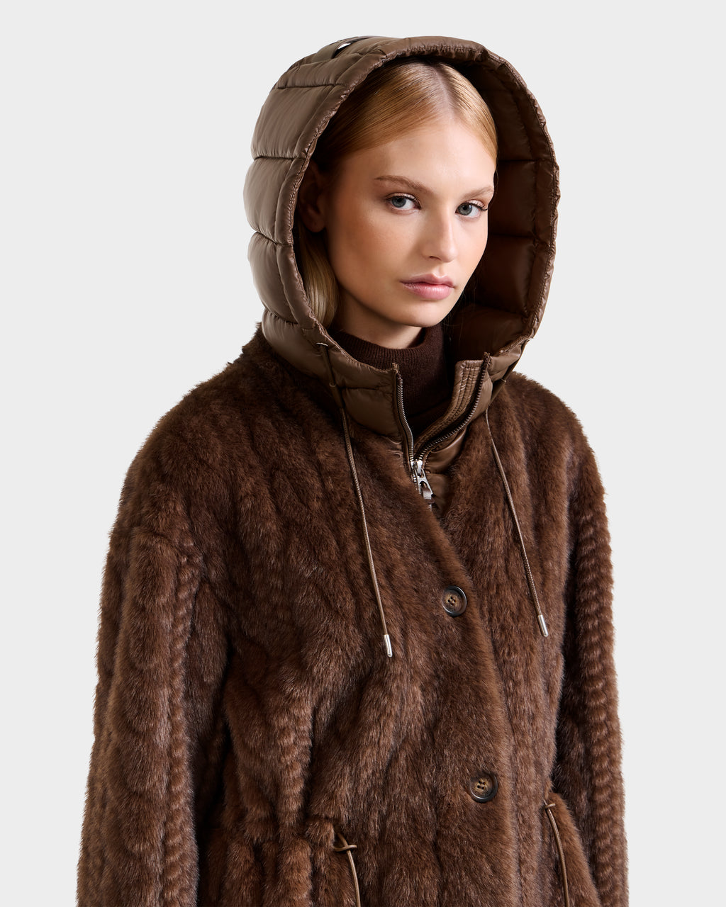 A woman wears the Rudsak ORIANA - 8125545 Mocha faux fur coat with a quilted hood and vegan leather trims, glancing to the side against a plain light background.