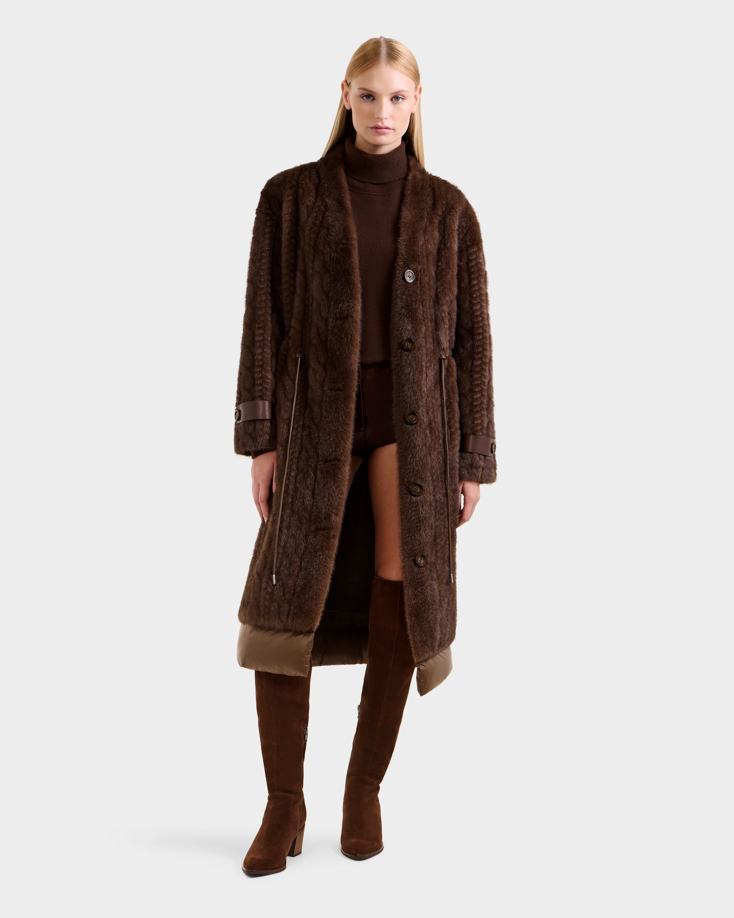 A woman with long blonde hair models the Rudsak ORIANA - 8125545 Mocha women’s faux fur coat with vegan leather trims, styled over a brown turtleneck, brown shorts, and knee-high suede boots against a plain light background.