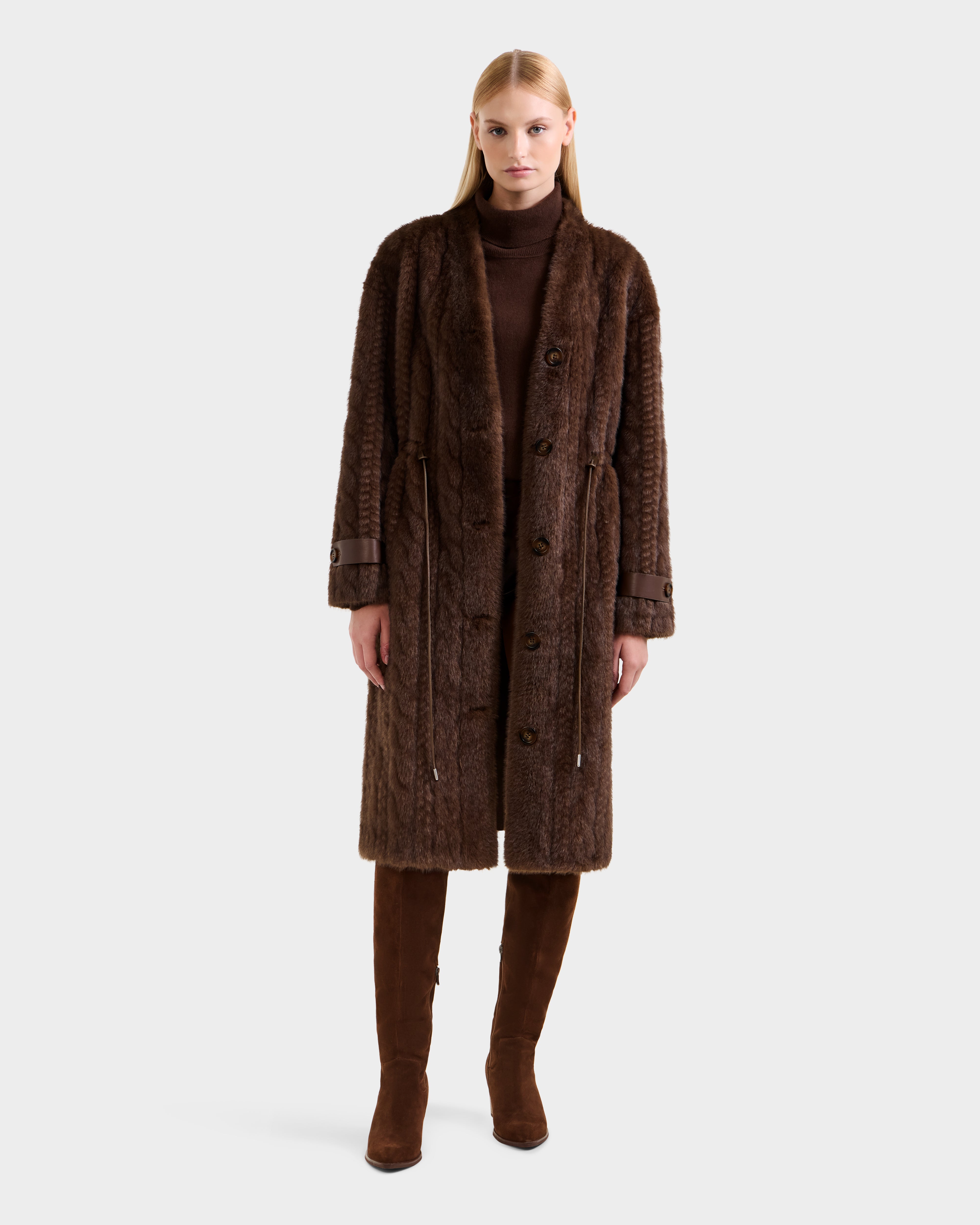 A woman with straight blonde hair wears the Rudsak ORIANA - 8125545 Mocha women’s faux fur coat with vegan leather trims over a brown turtleneck and knee-high boots, standing against a light gray background.