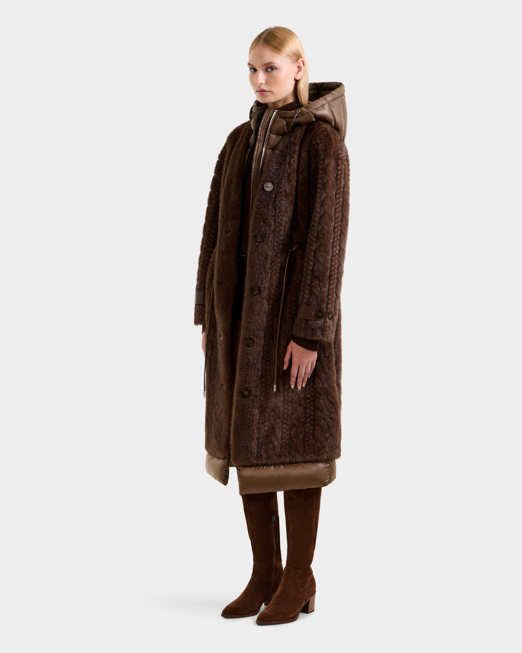 A woman stands facing forward in the ORIANA - 8125545 Mocha coat by Rudsak, featuring vegan leather trims. She pairs it with brown knee-high boots, her arms relaxed at her sides and straight blonde hair, set against a plain light grey background.