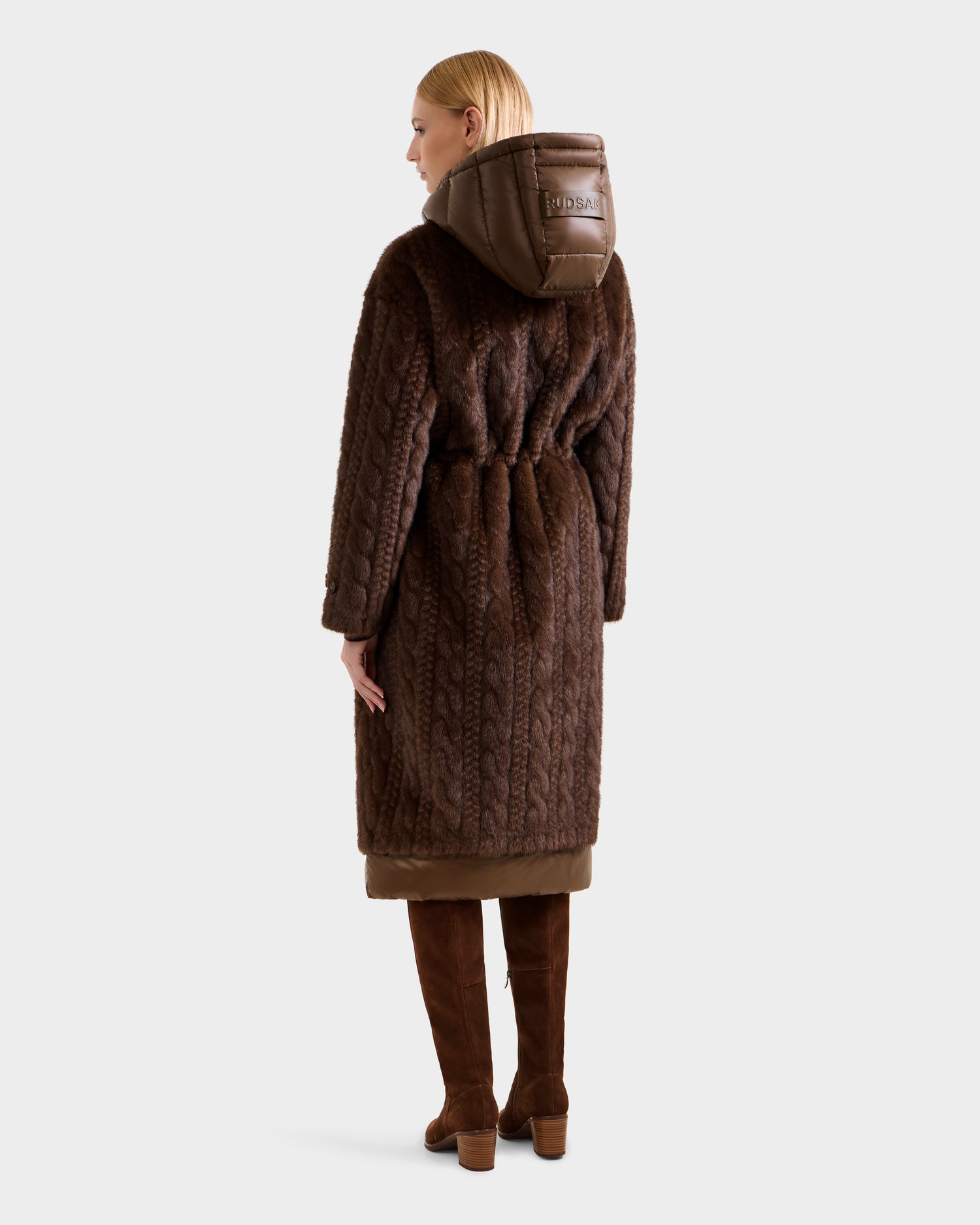 A person stands facing away, wearing the Rudsak ORIANA - 8125545 Mocha women’s faux fur coat with a hood, vegan leather trims, waist belt, and logo patch on the hood. Matching knee-high boots complete the look against a plain background.