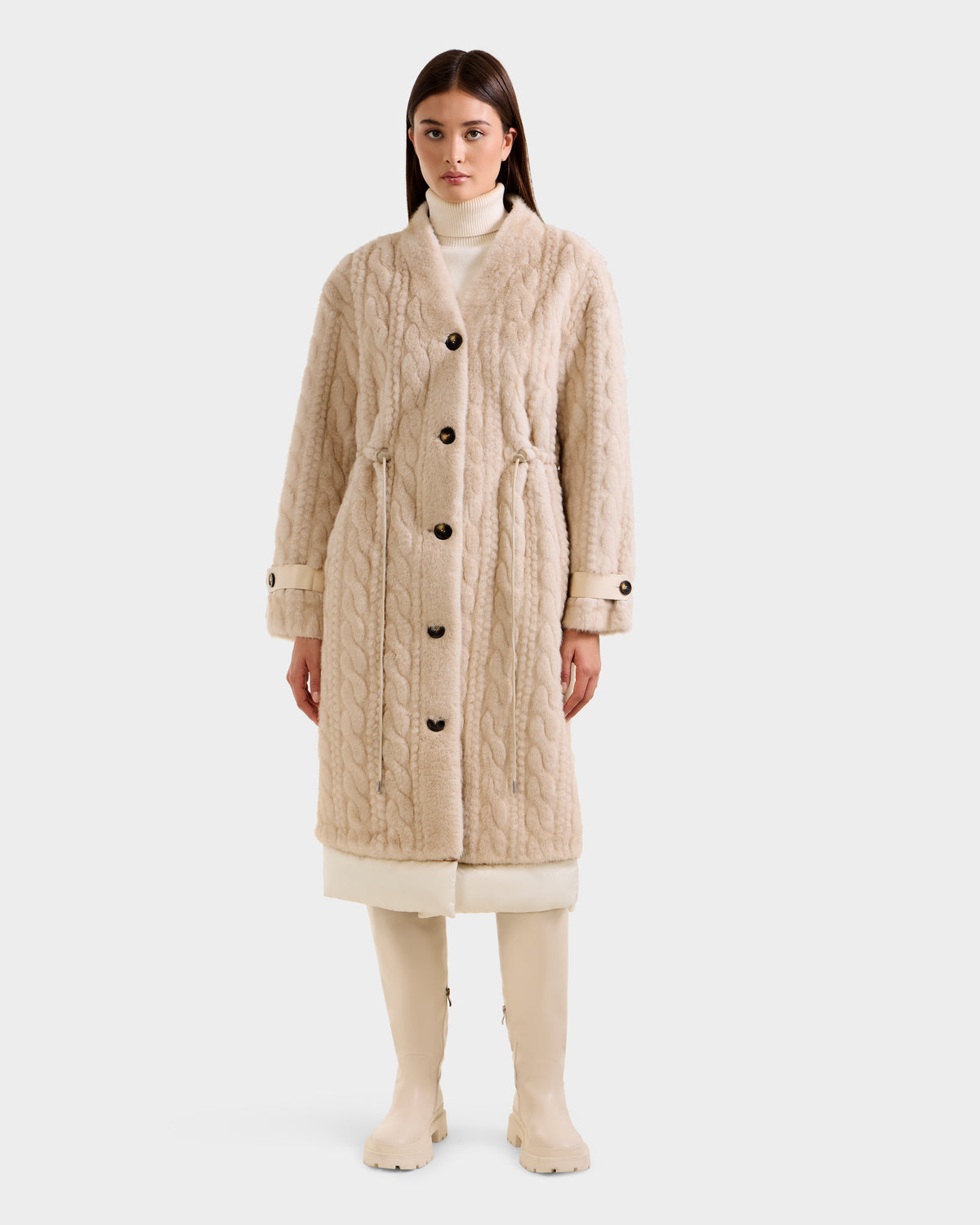 A woman faces forward against a plain light background, wearing the Rudsak ORIANA - 8125545 Vanilla cream cable knit coat with large buttons and vegan leather trim, styled with a high-neck white top and knee-high cream boots.