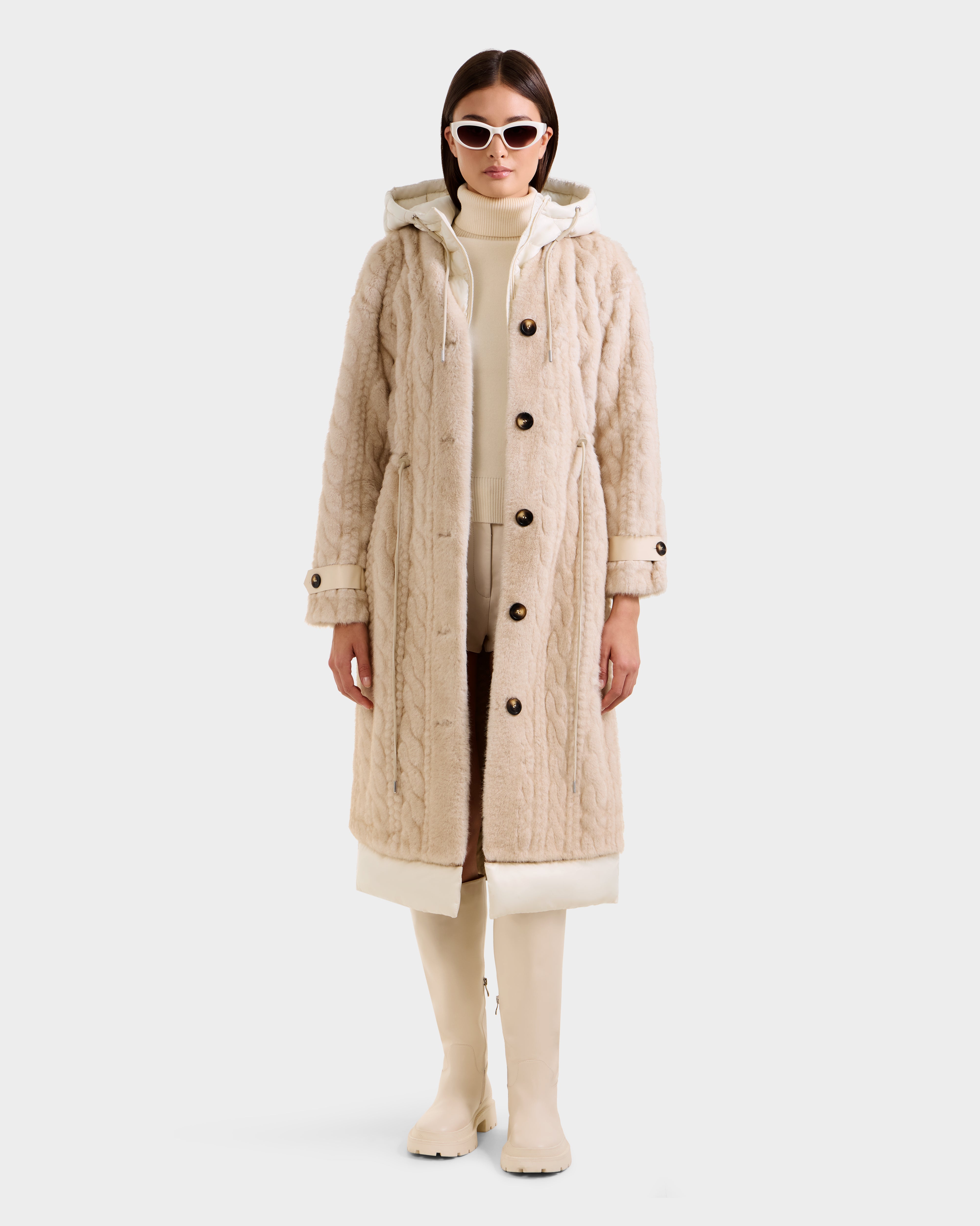 A woman wears the Rudsak ORIANA - 8125545 Vanilla cable knit coat with buttons and hood, styled with a cream turtleneck, light pants, knee-high boots, and white sunglasses against a plain white background.