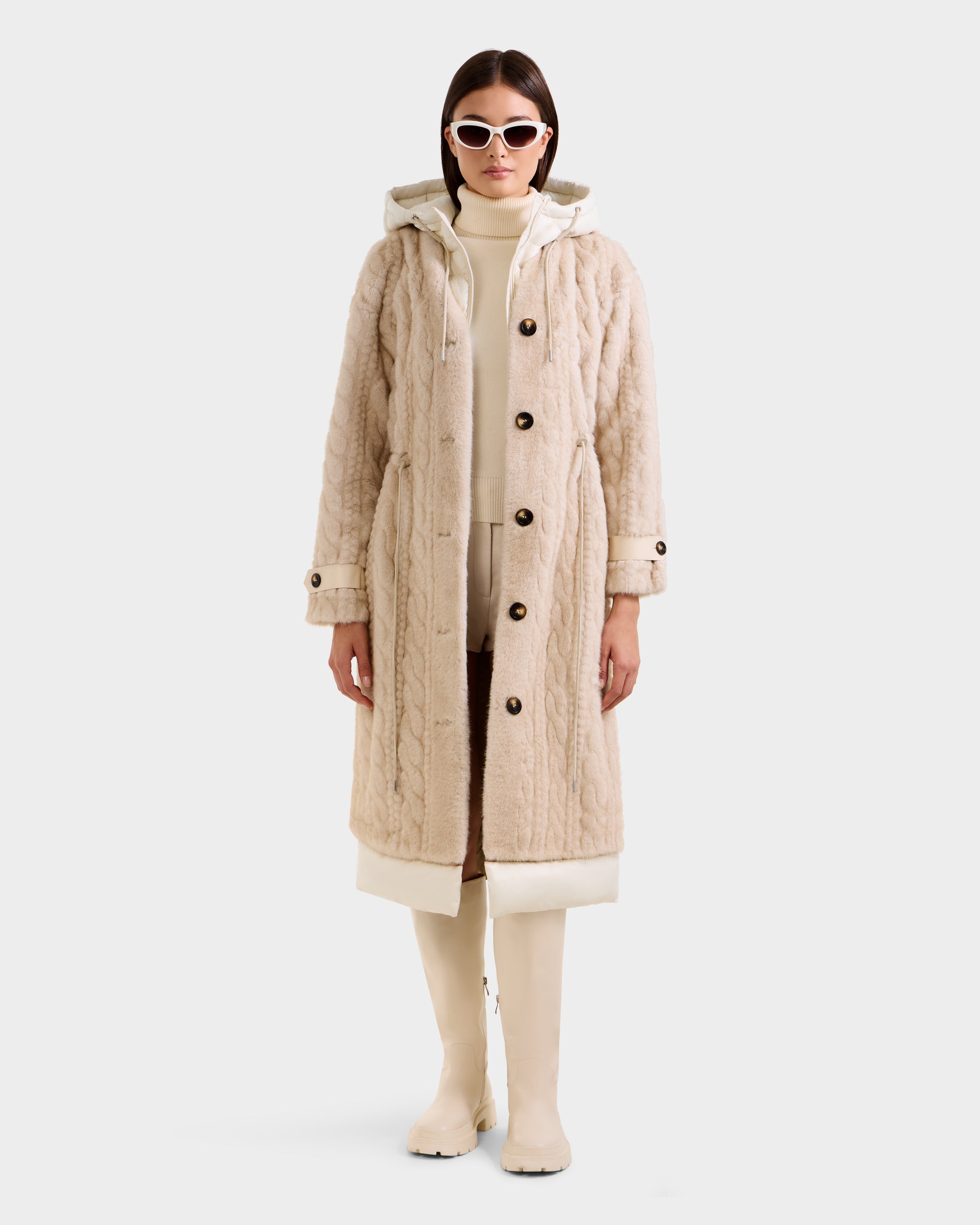 A woman wears the Rudsak ORIANA - 8125545 Vanilla cable knit coat with buttons and hood, styled with a cream turtleneck, light pants, knee-high boots, and white sunglasses against a plain white background.
