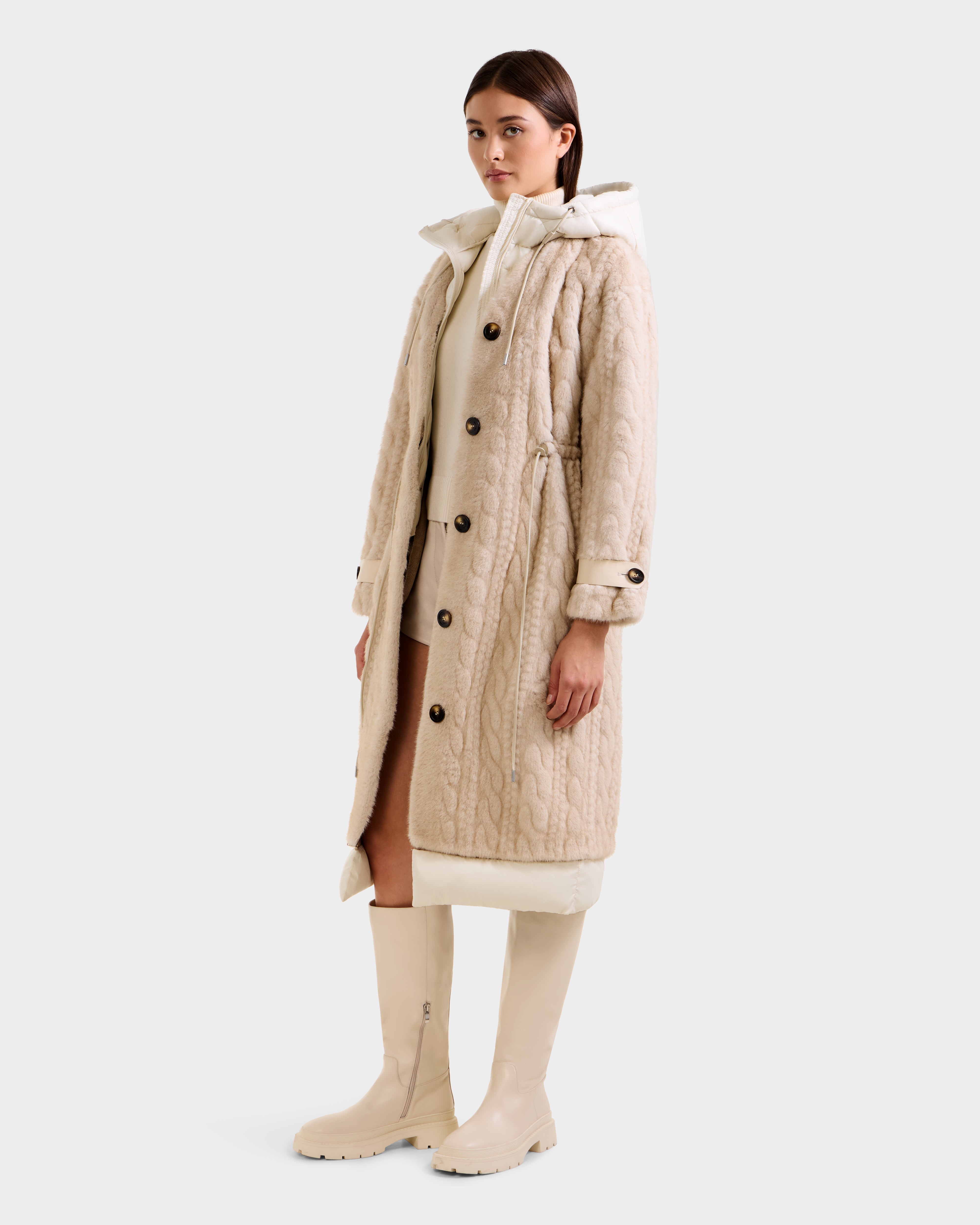 A woman wears the Rudsak ORIANA - 8125545 Vanilla, a long cream quilted coat with vegan leather trim and large buttons over a white hooded layer and beige knee-high boots, standing slightly turned against a plain light background.