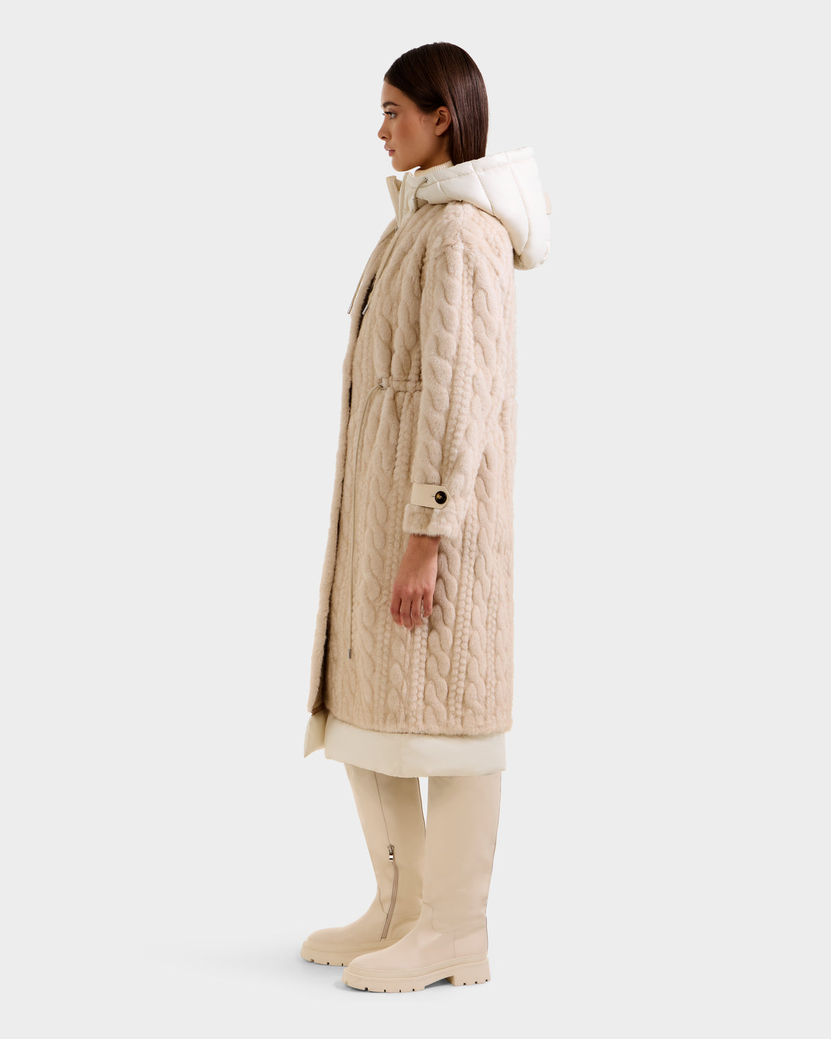 A woman stands in profile against a plain light background, wearing the Rudsak ORIANA - 8125545 Vanilla long textured beige coat with vegan leather trim, paired with cream leggings and knee-high beige boots.