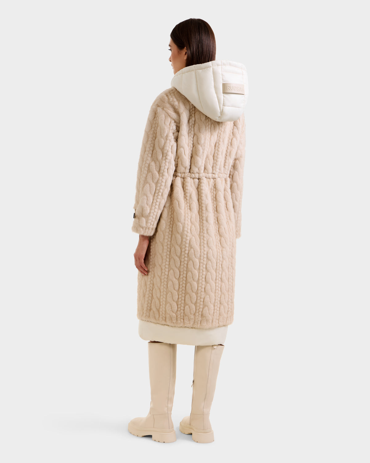 A woman faces away wearing the Rudsak ORIANA - 8125545 Vanilla, a long beige cable knit coat with a white quilted hood and matching tall beige boots, against a plain white background.