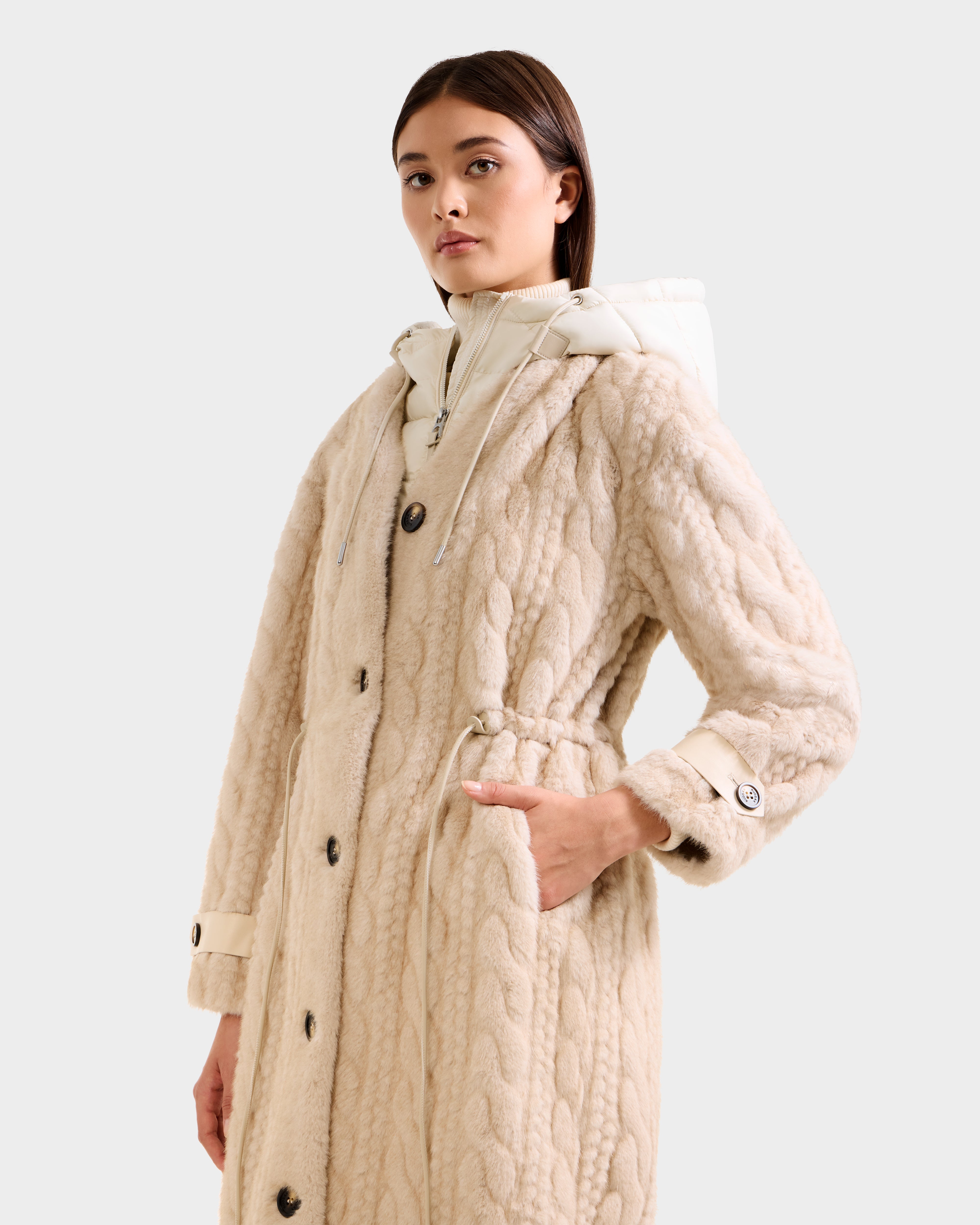 A woman models the Rudsak ORIANA - 8125545 Vanilla, a long textured beige coat with large buttons and a drawstring waist, standing with one hand in her pocket against a plain light background.