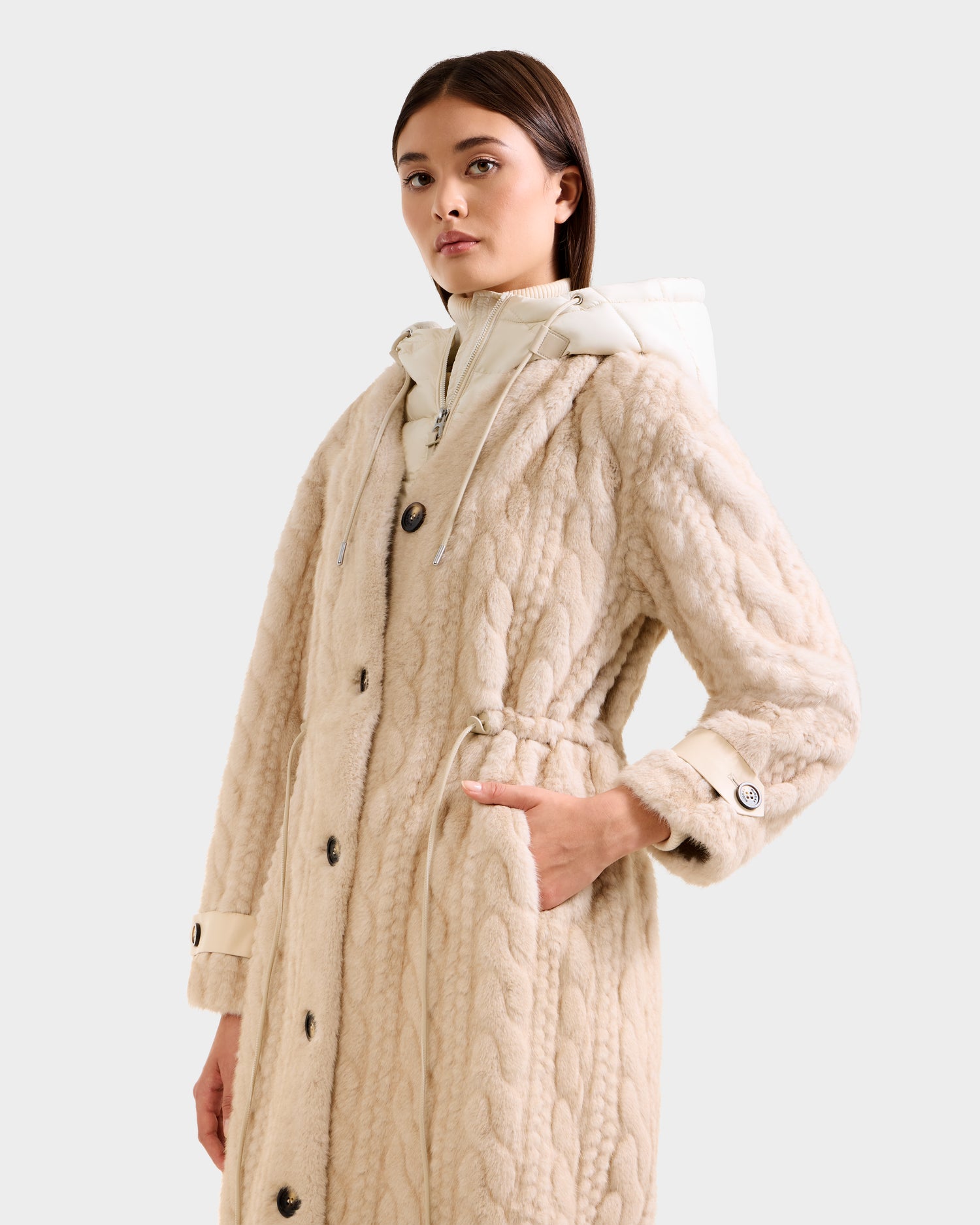 A woman models the Rudsak ORIANA - 8125545 Vanilla, a long textured beige coat with large buttons and a drawstring waist, standing with one hand in her pocket against a plain light background.