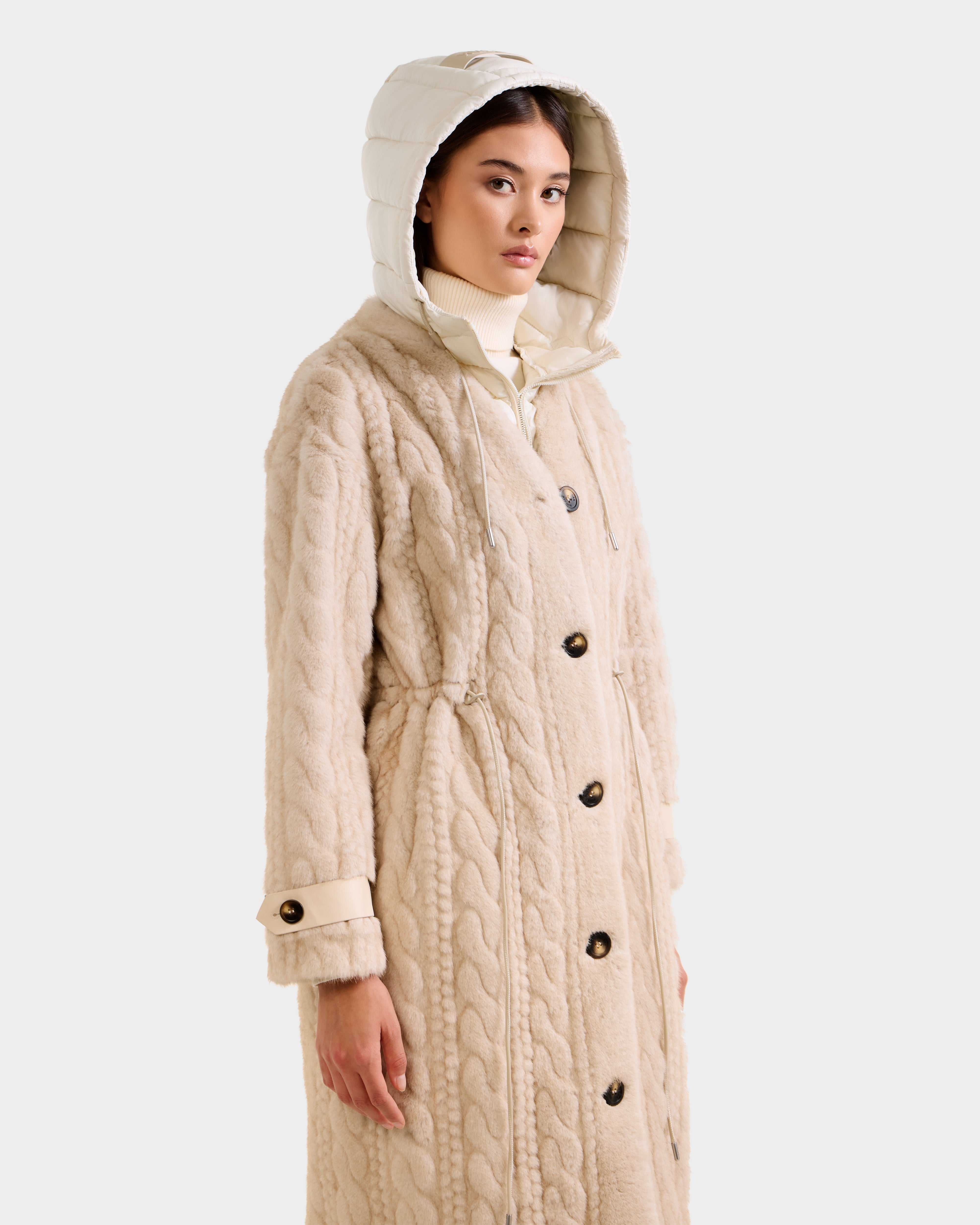 A woman wearing the Rudsak ORIANA - 8125545 Vanilla, a long beige coat with large buttons, vegan leather trim, and padded hood, stands against a plain light background, looking slightly to the side.