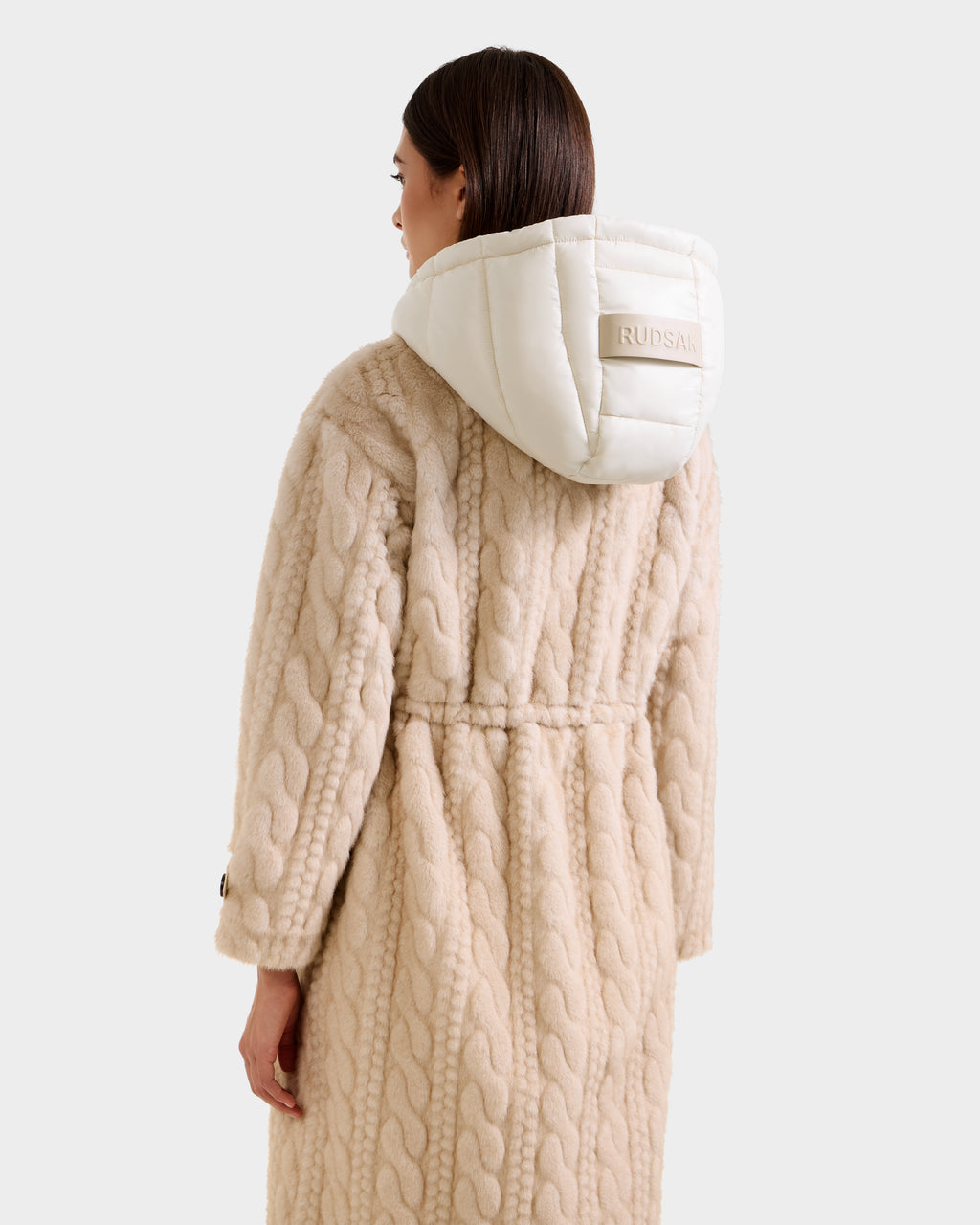 A woman is shown from the back wearing the Rudsak ORIANA - 8125545 Vanilla, a long textured beige cable knit coat with a large white hood featuring a patch. The background is plain and light-colored.