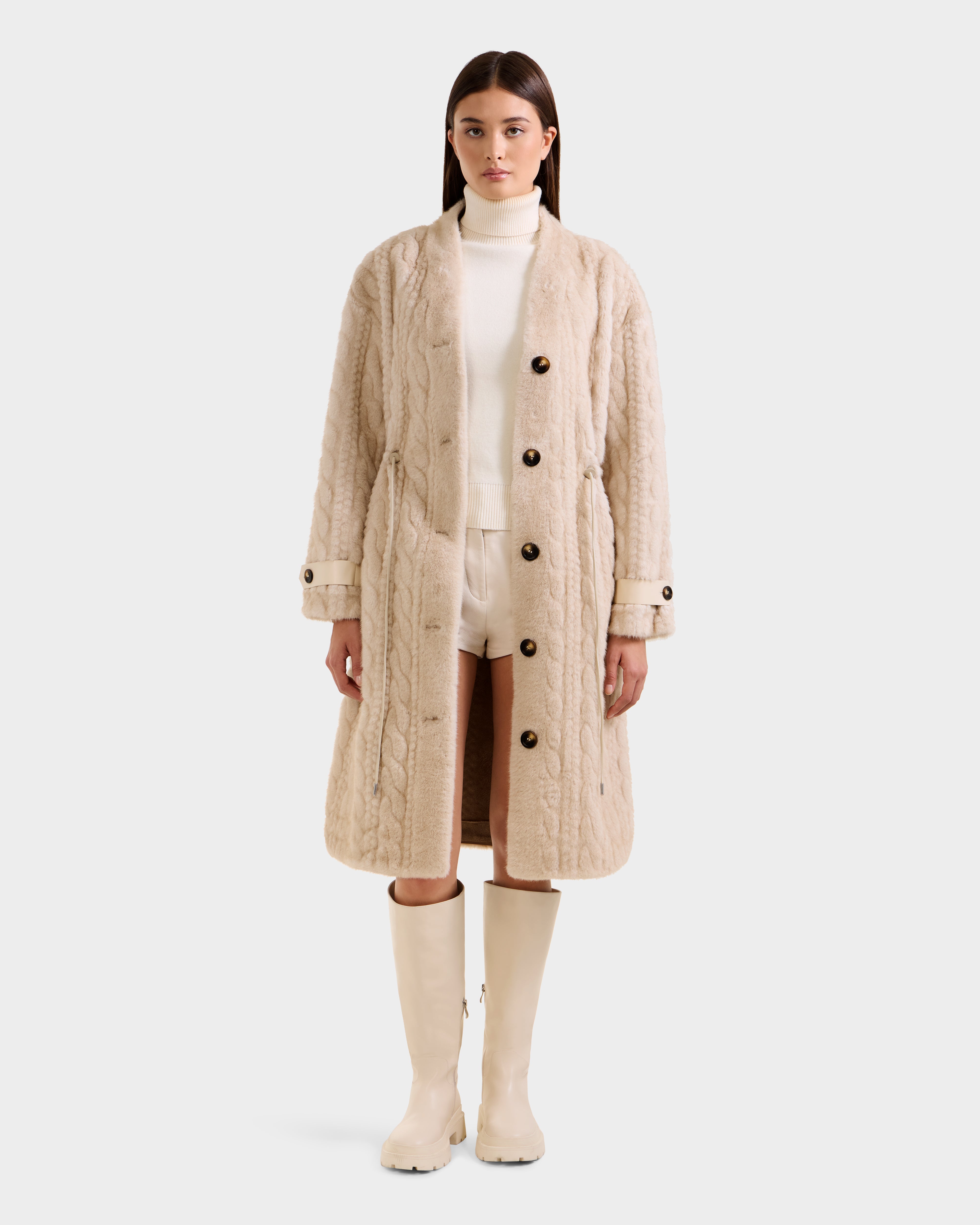 A woman faces forward wearing the Rudsak ORIANA - 8125545 Vanilla cable knit coat with large buttons, styled with a white turtleneck, white shorts, and cream knee-high boots against a plain light background.