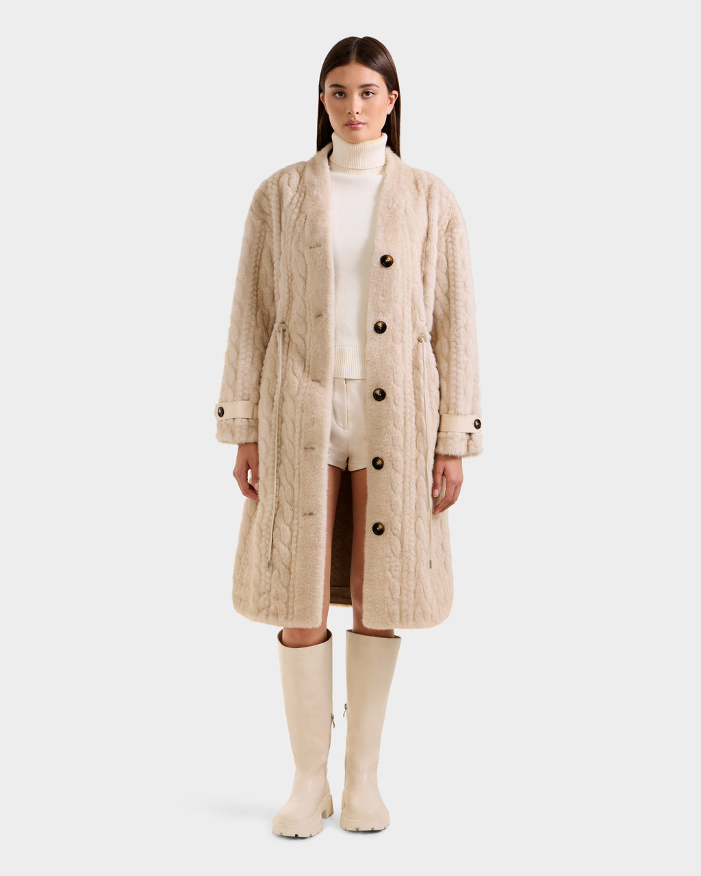 A woman faces forward wearing the Rudsak ORIANA - 8125545 Vanilla cable knit coat with large buttons, styled with a white turtleneck, white shorts, and cream knee-high boots against a plain light background.