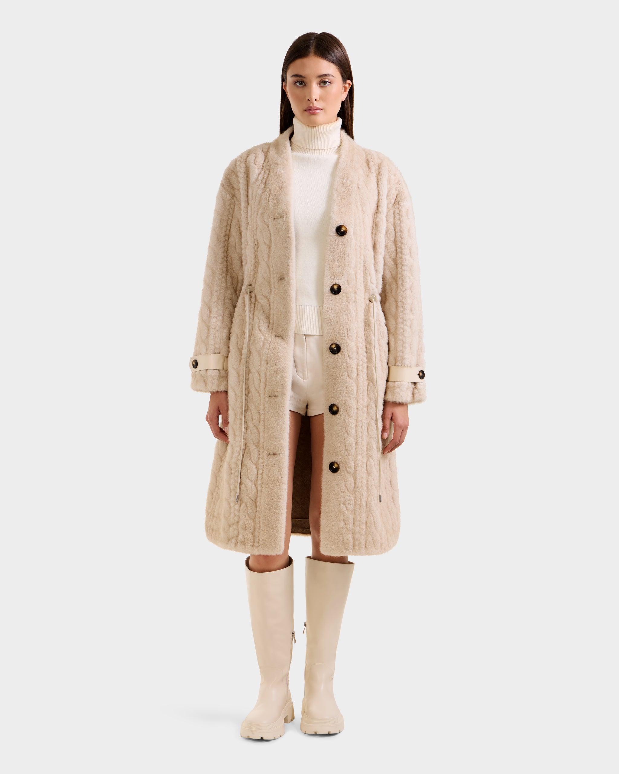 A woman faces forward wearing the Rudsak ORIANA - 8125545 Vanilla cable knit coat with large buttons, styled with a white turtleneck, white shorts, and cream knee-high boots against a plain light background.