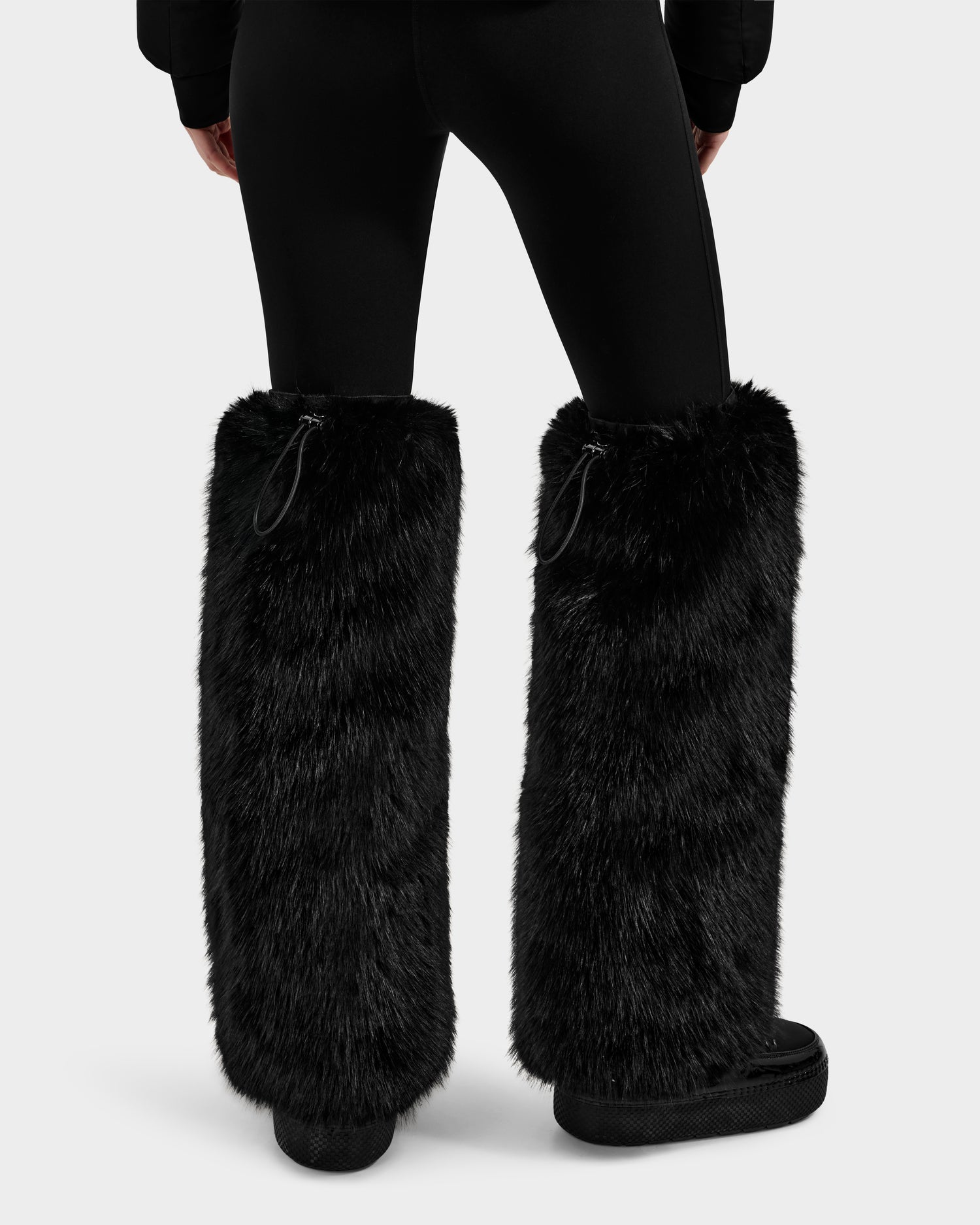 A person showcases après-ski style, wearing Rudsak PAIGE - 8425534 Black faux fur leg warmers over black leggings and shoes, viewed from the back against a plain light background.