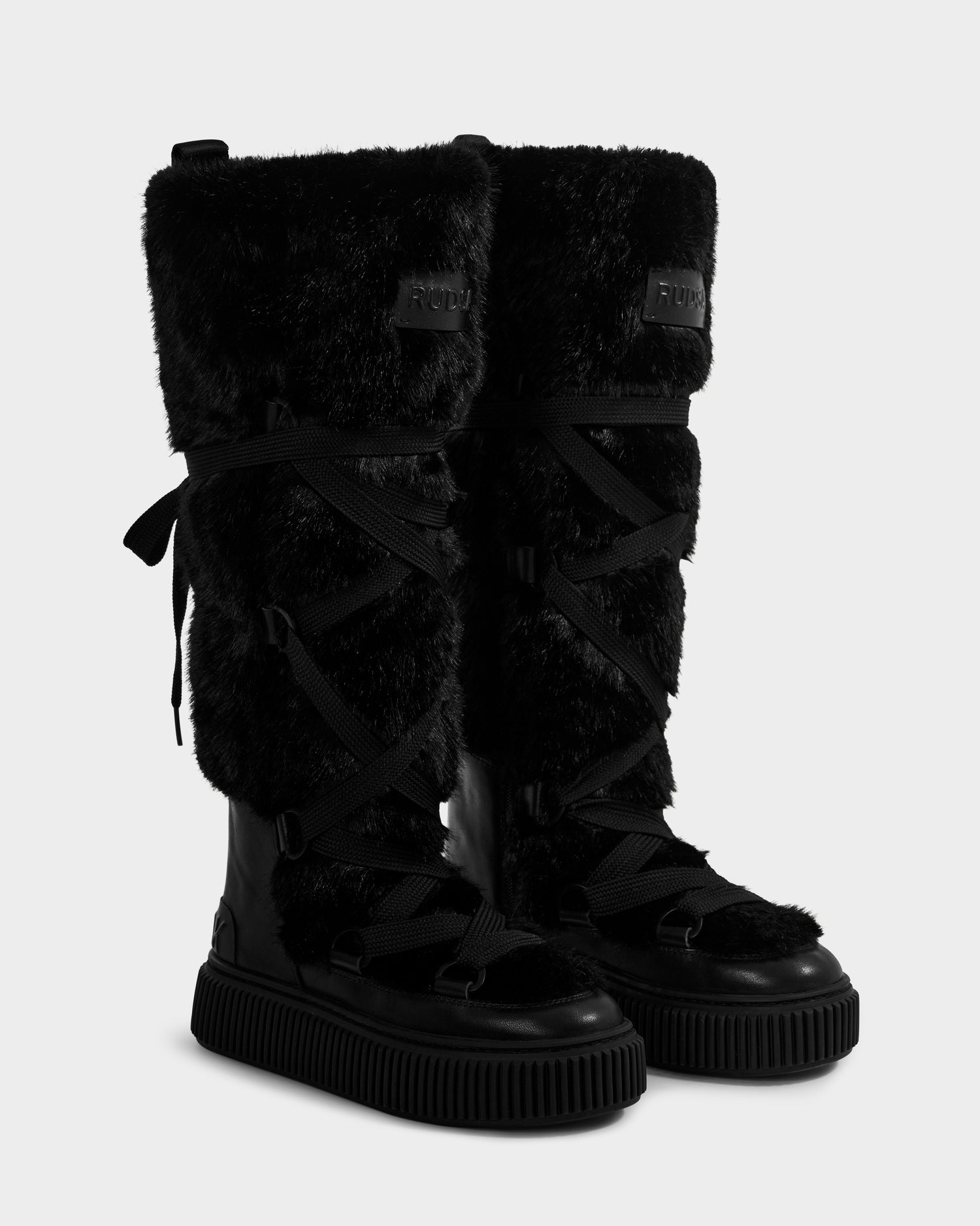 The Rudsak PALERMO C - 8225546 Black women’s boots feature tall black faux fur, crisscross laces, and ribbed rubber soles, displayed against a plain light background.