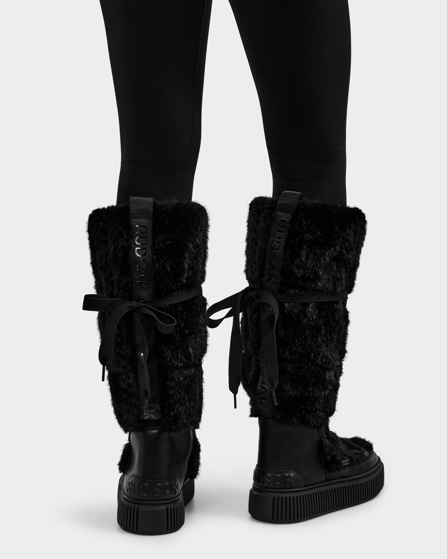 A person wearing black leggings and tall black Rudsak PALERMO C - 8225546 women’s faux fur winter boots with thick soles, ribbon laces tied in bows at the back, and a visible Rudsak label on the footwear.
