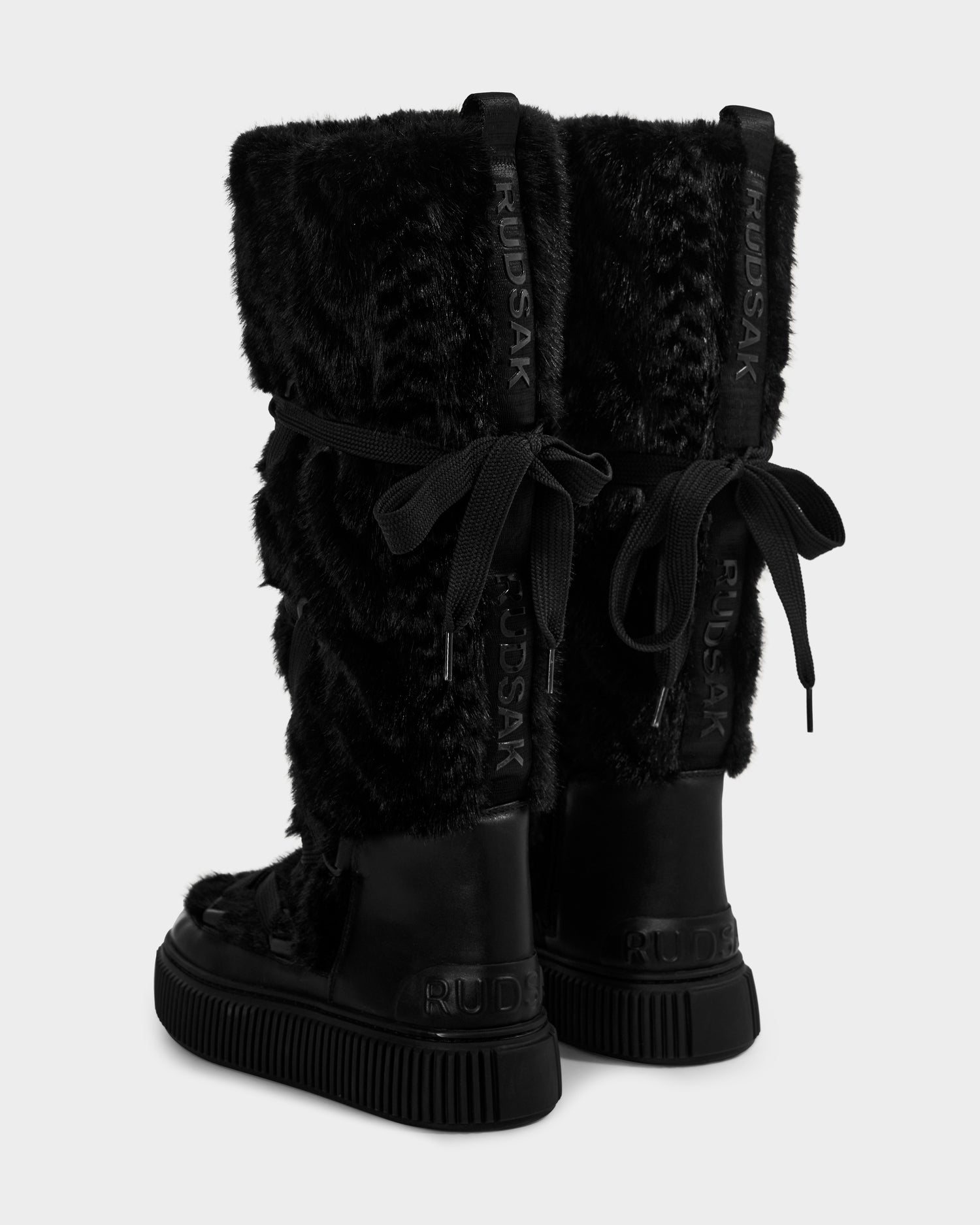 The PALERMO C - 8225546 Black by Rudsak are black knee-high women’s faux fur winter boots with thick soles, ribbon laces at the back, and Rudsak branding on the heel and back seam—ideal for winter.