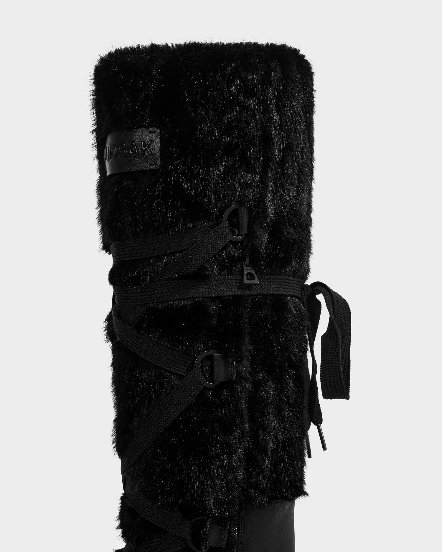 A close-up of the Rudsak PALERMO C - 8225546 Black women’s après-ski boot features a faux fur exterior, crisscrossed laces, and a fabric tag, highlighting its texture and style against a plain white background.
