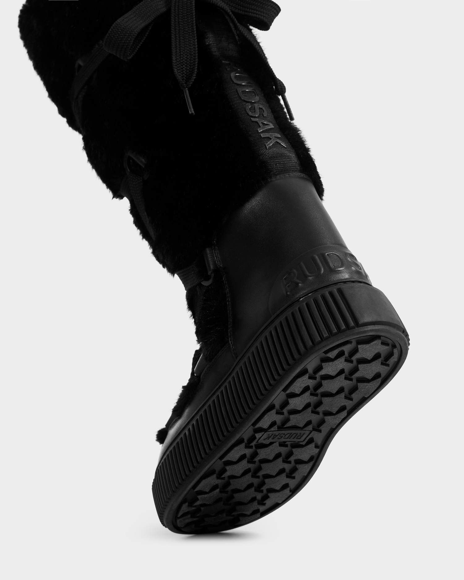 Close-up of the Rudsak PALERMO C - 8225546 Black women’s faux fur winter boot with laces, a textured rubber sole, and RUDSAK embossed on the side and heel. The worn boot is slightly lifted at the heel.