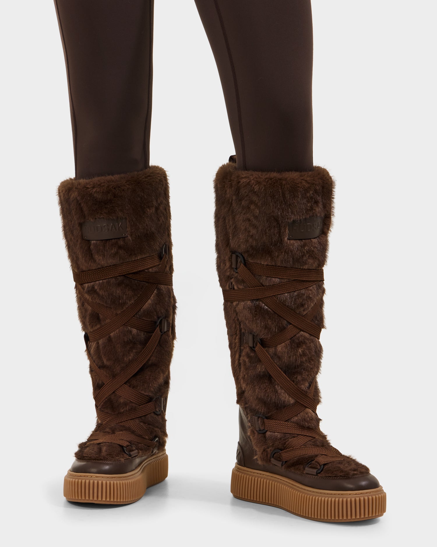 A person wears Rudsak PALERMO C - 8225546 Mocha knee-high women’s faux fur winter boots with crisscrossed brown laces and thick rubber soles, paired with dark brown leggings—a perfect cozy winter look.