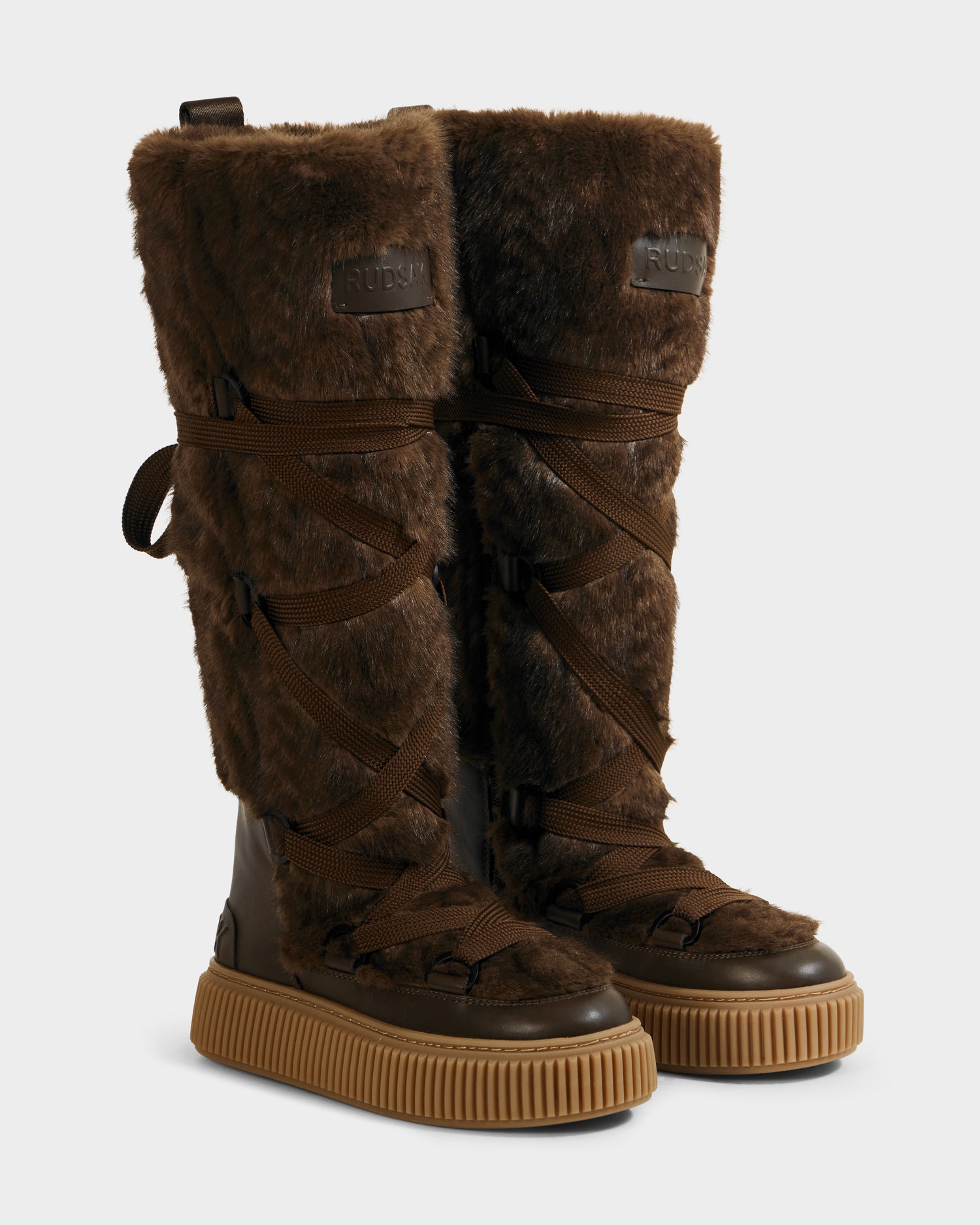 The Rudsak PALERMO C - 8225546 Mocha is a pair of tall women’s faux fur winter boots in brown, featuring lace-up fronts, rubber soles, woven fabric with cable knit pattern, and a RUBY-embossed patch on the upper front.