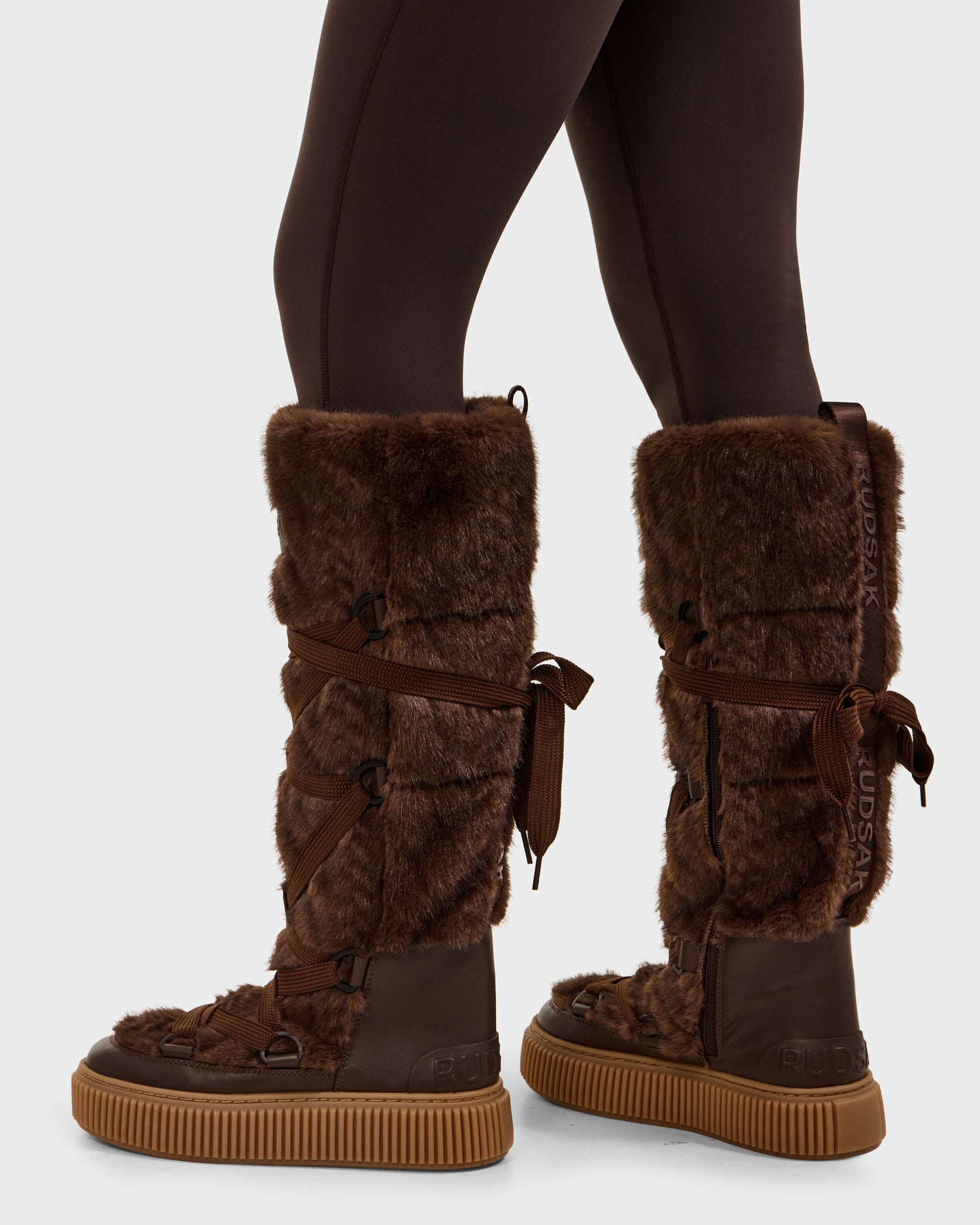 A person wearing Rudsak PALERMO C - 8225546 Mocha women’s faux fur winter boots with thick rubber soles and ribbon laces, paired with dark leggings—the perfect après-ski look—shown from the side and back.