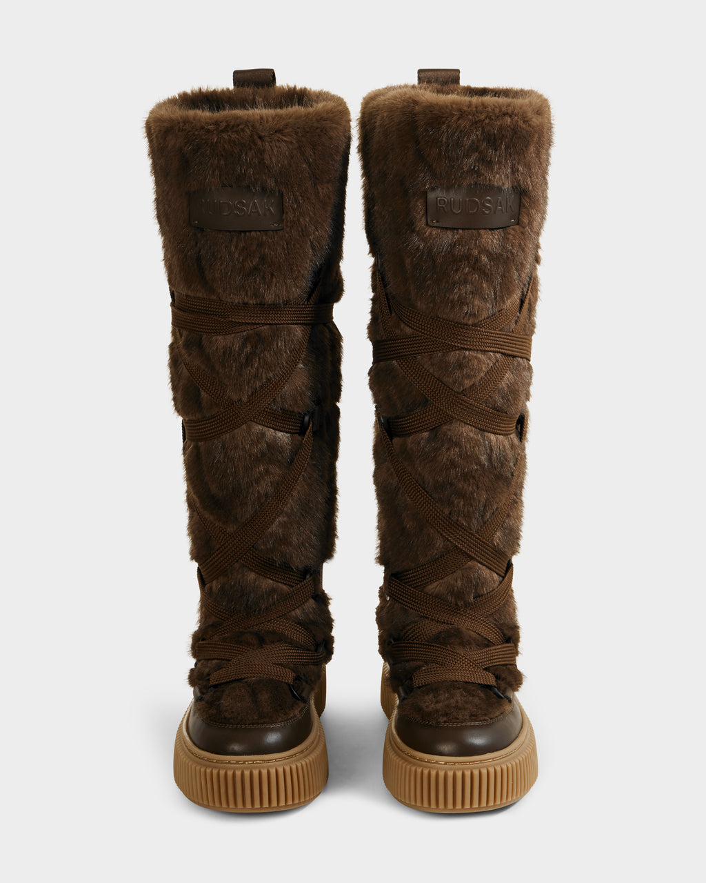 The PALERMO C - 8225546 Mocha by Rudsak are knee-high women’s faux fur winter boots with crisscross dark brown laces, rubber soles, subtle cable knit detail, and a Rudsak leather patch at the top front.