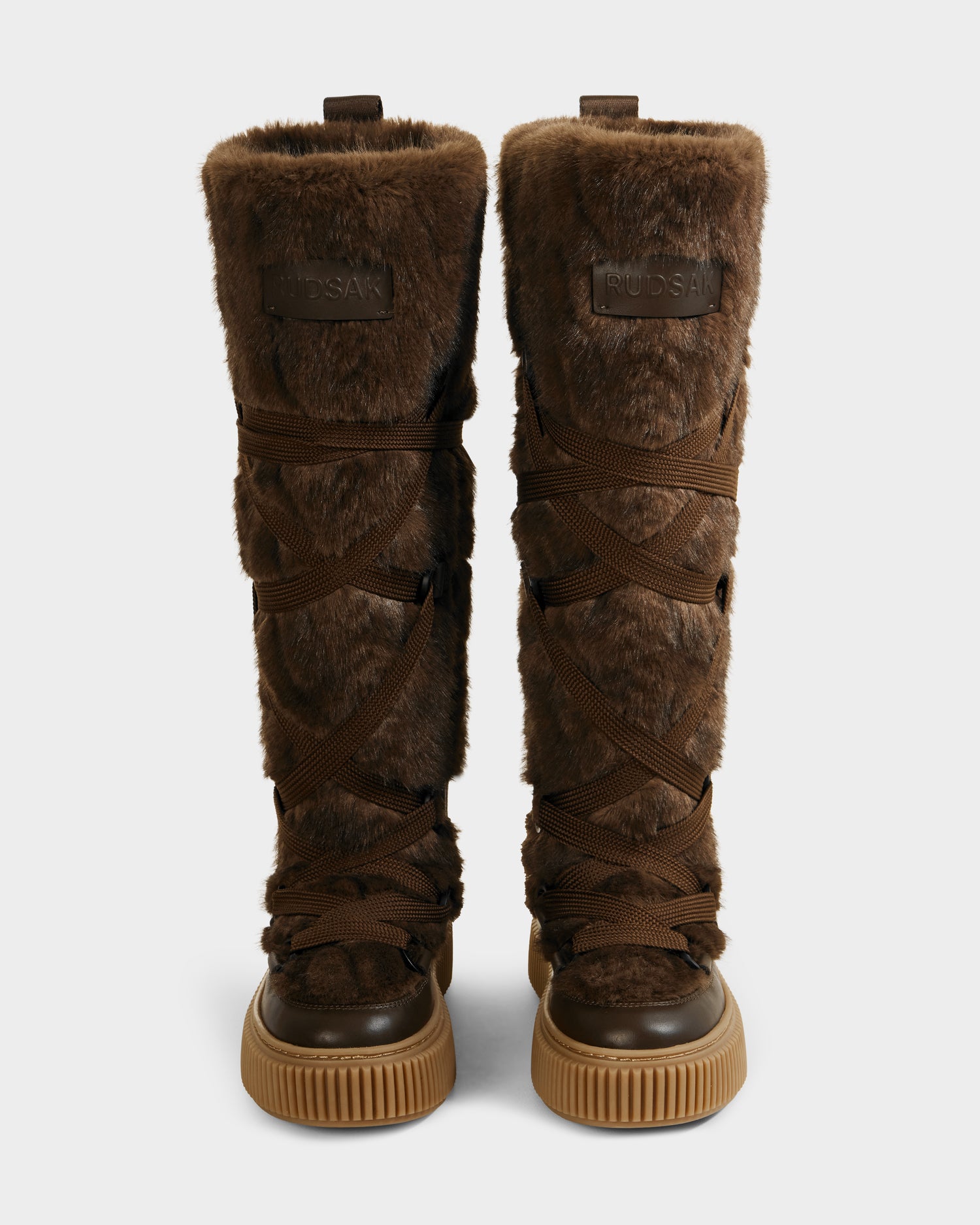 The PALERMO C - 8225546 Mocha by Rudsak are knee-high women’s faux fur winter boots with crisscross dark brown laces, rubber soles, subtle cable knit detail, and a Rudsak leather patch at the top front.