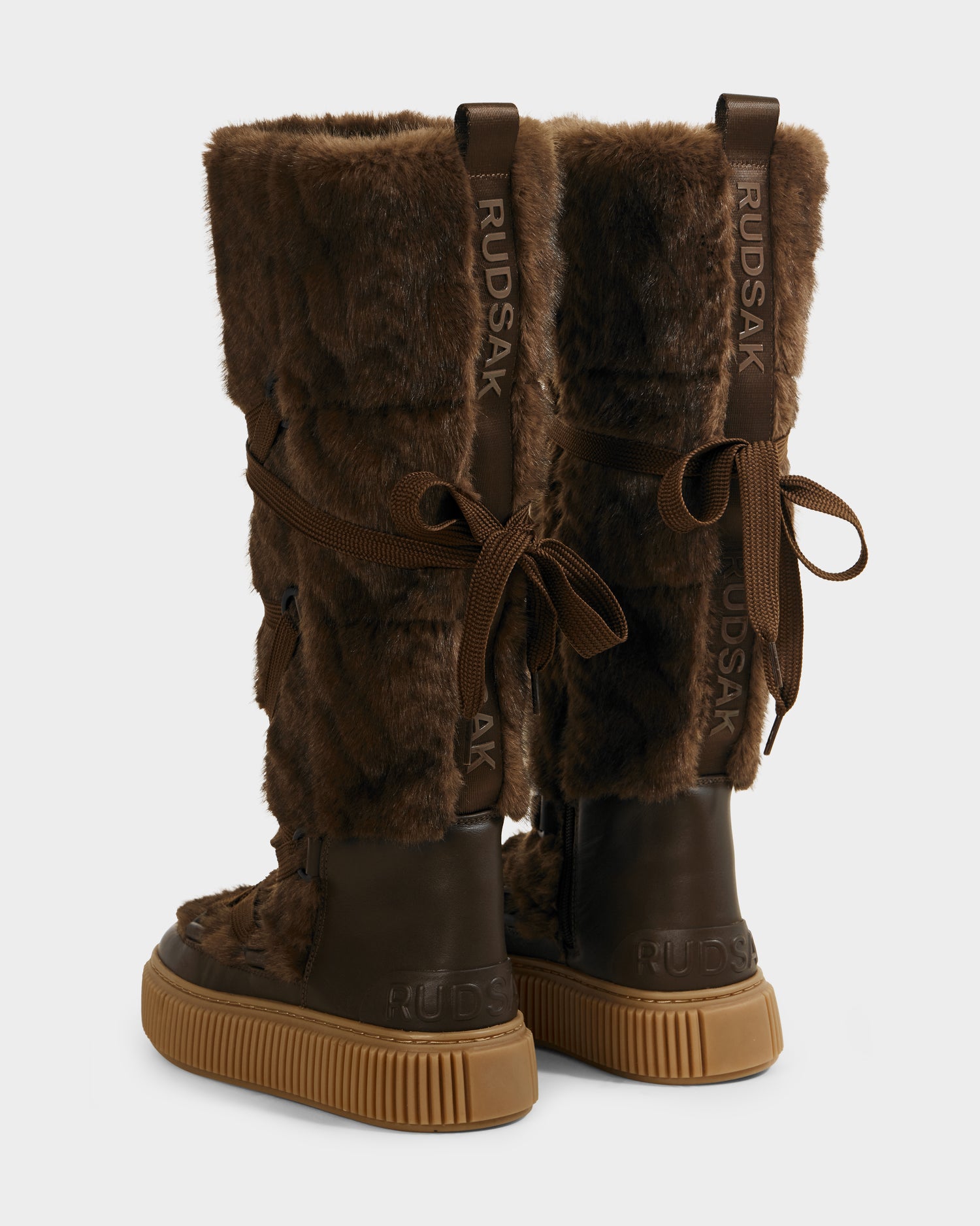 The PALERMO C - 8225546 Mocha by Rudsak are tall women’s mocha faux fur winter boots featuring brown rubber soles, back-tied laces, and "RUDSAK" printed vertically on the rear. Shown from a rear angle for style emphasis.