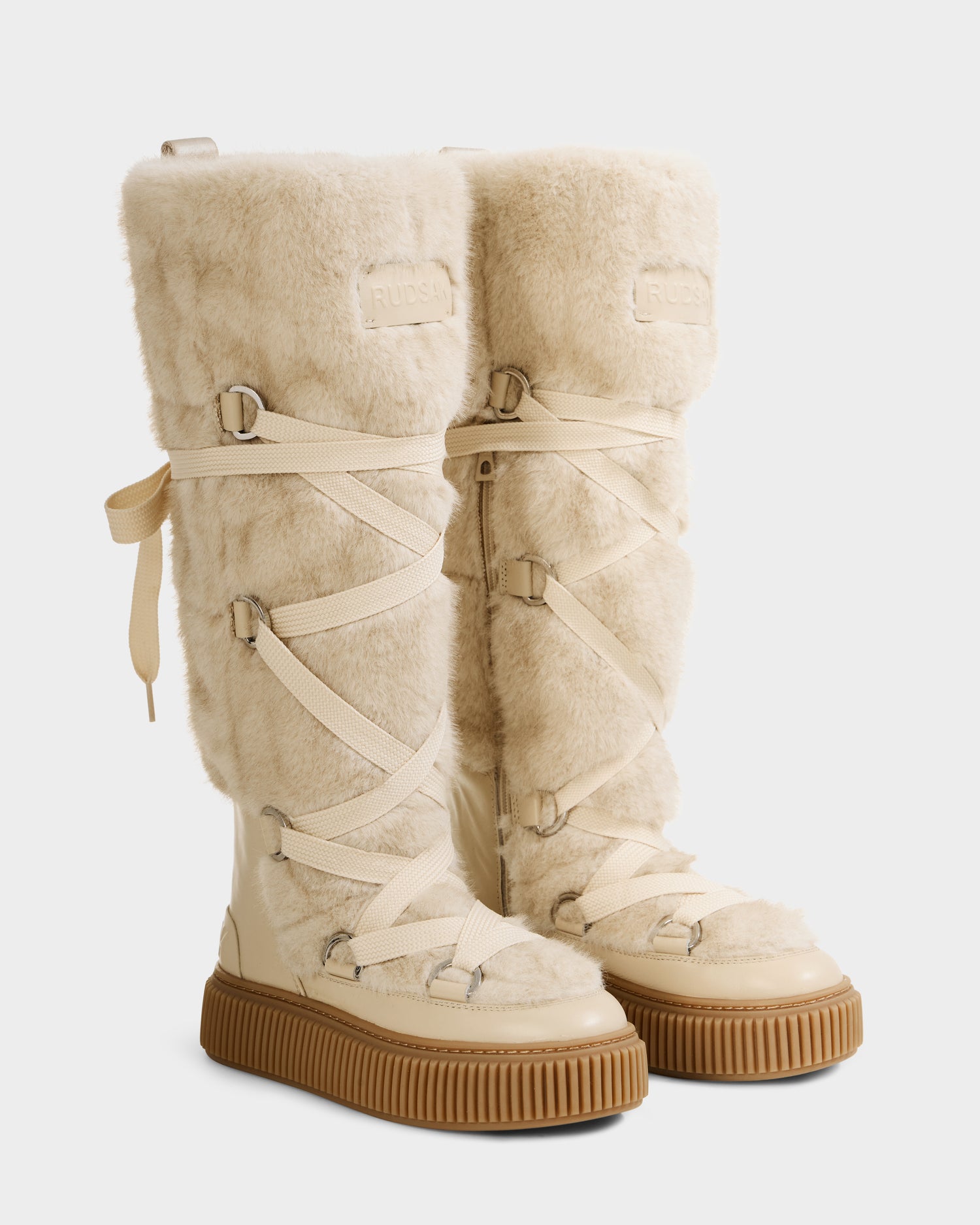 The Rudsak PALERMO C - 8225546 Vanilla boots are beige, knee-high women's winter boots with cozy faux fur uppers, ribbed tan rubber soles, cream crisscross laces, and an embossed logo—ideal for chic après-ski style.
