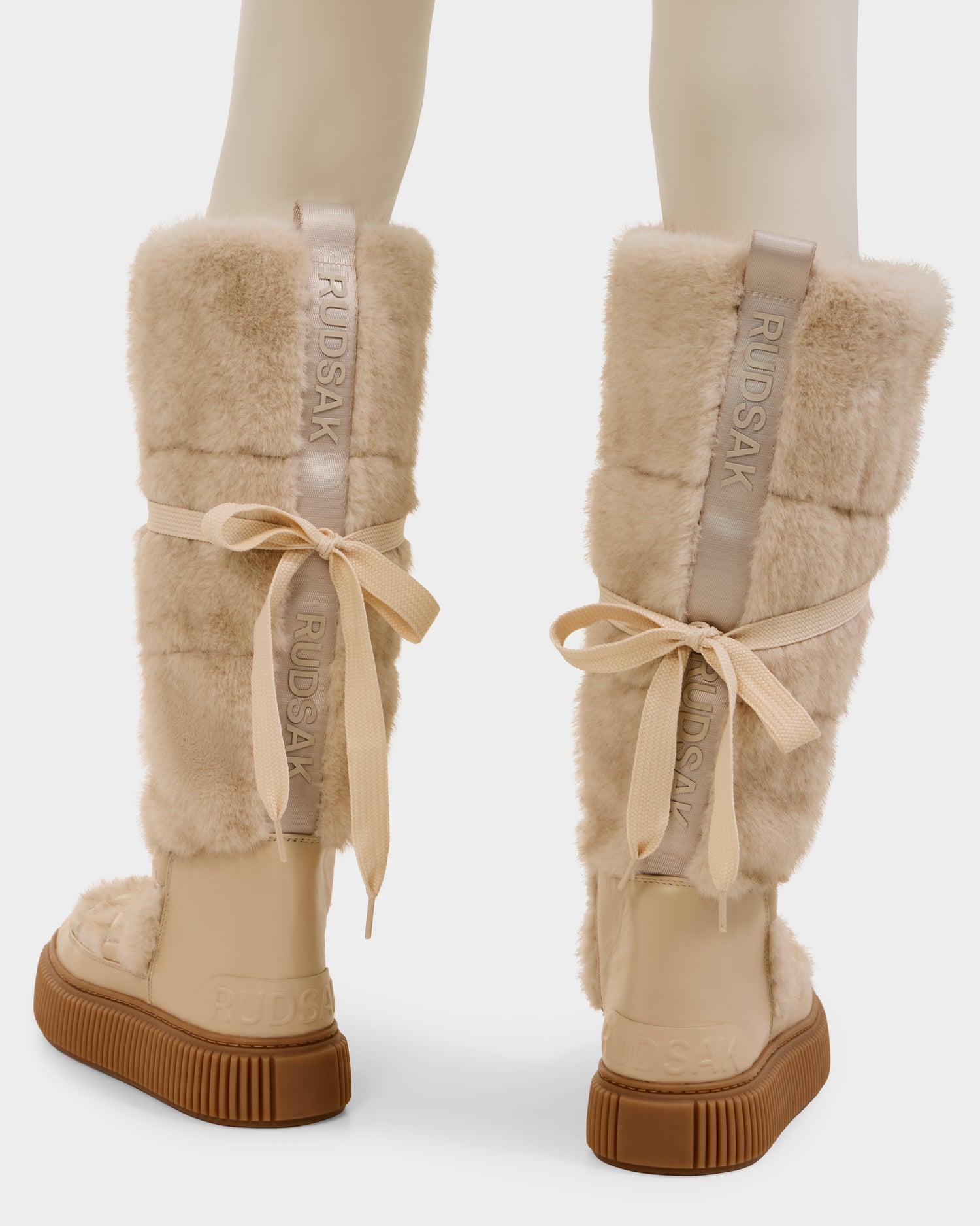 A pair of beige, knee-high women’s winter boots—PALERMO C - 8225546 Vanilla by Rudsak—feature faux fur, ribbon laces, thick rubber soles, and Rudsak branding on fabric strips down the back. Shown from the rear view.