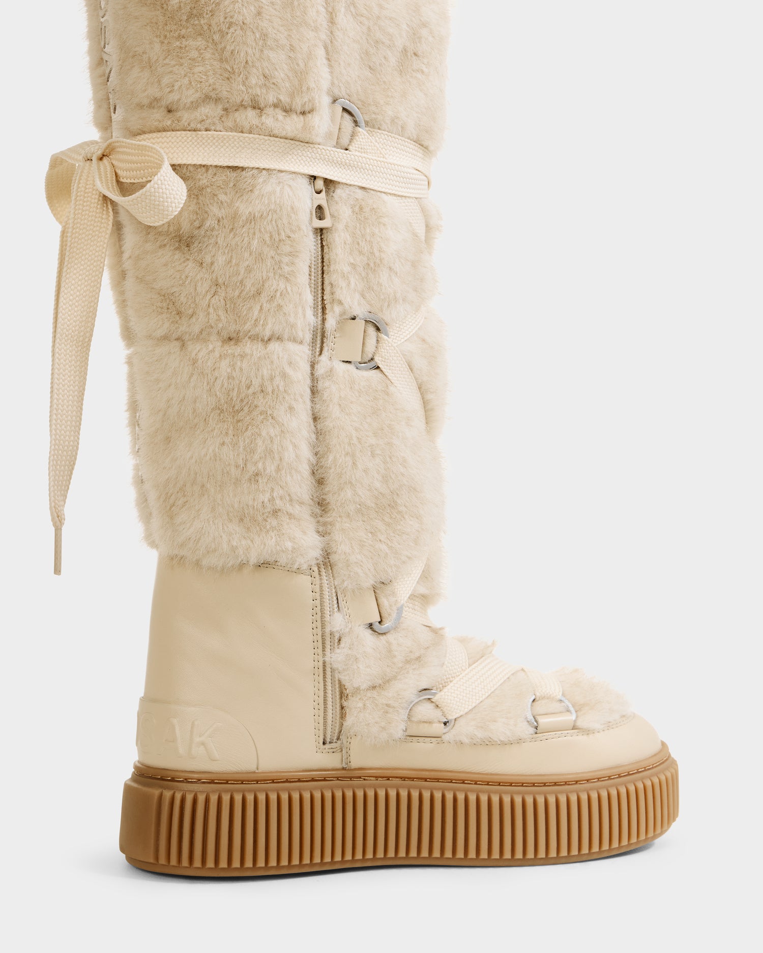 A close-up side view of the Rudsak PALERMO C - 8225546 Vanilla women’s faux fur winter boot in beige, featuring cream laces, metal hardware, a thick brown rubber sole, textured details, visible zippers, and a decorative side bow.