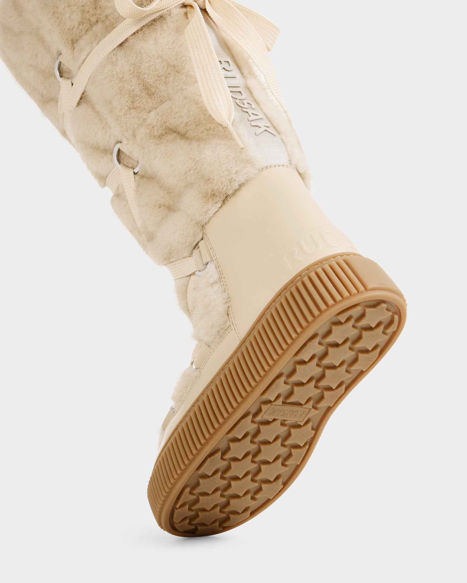 Close-up of a person wearing Rudsak PALERMO C - 8225546 Vanilla women’s faux fur winter boots with a beige rubber sole and crisscrossed laces, lifting their foot to display the sole’s texture—stylish winter footwear on a white background.