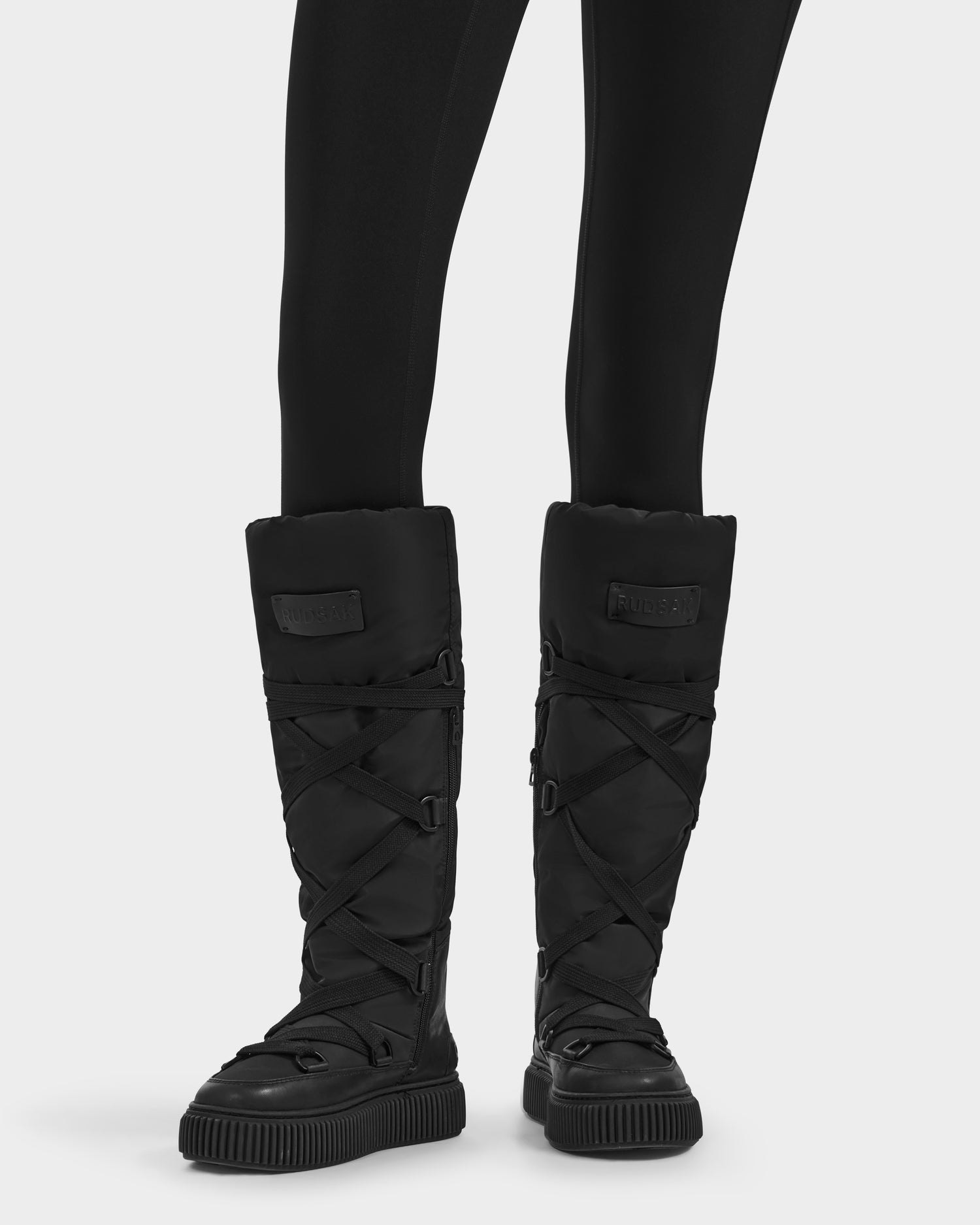 A person wearing Rudsak's PALERMO - 8225030 Black insulated knee-high boots with crisscross straps and thick soles, paired with black leggings, stands against a plain light background.
