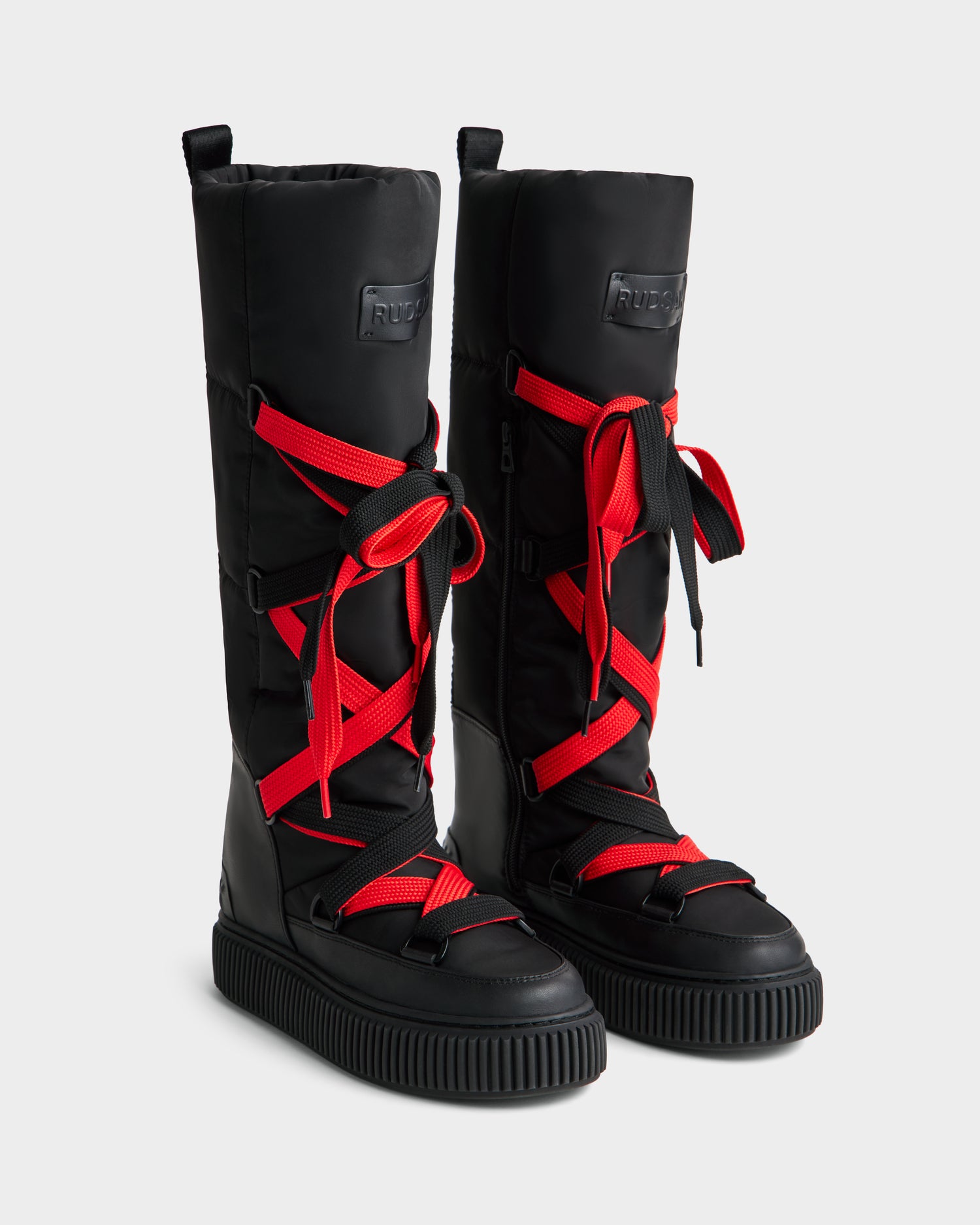 The Rudsak PALERMO - 8225030 Black boots are tall, insulated with chunky soles, bright red crisscross laces, and a rectangular logo patch on the front, shown against a plain light background.