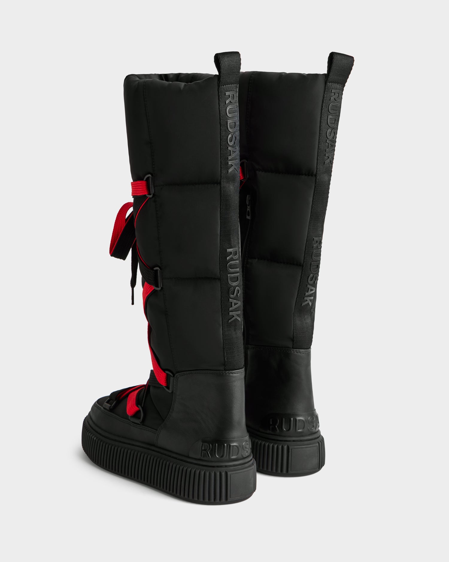 Two tall black PALERMO - 8225030 women’s winter boots by Rudsak feature thick soles, a water-resistant quilted design, red back laces, and RUDSAK embossed on the heel and vertically along the back.