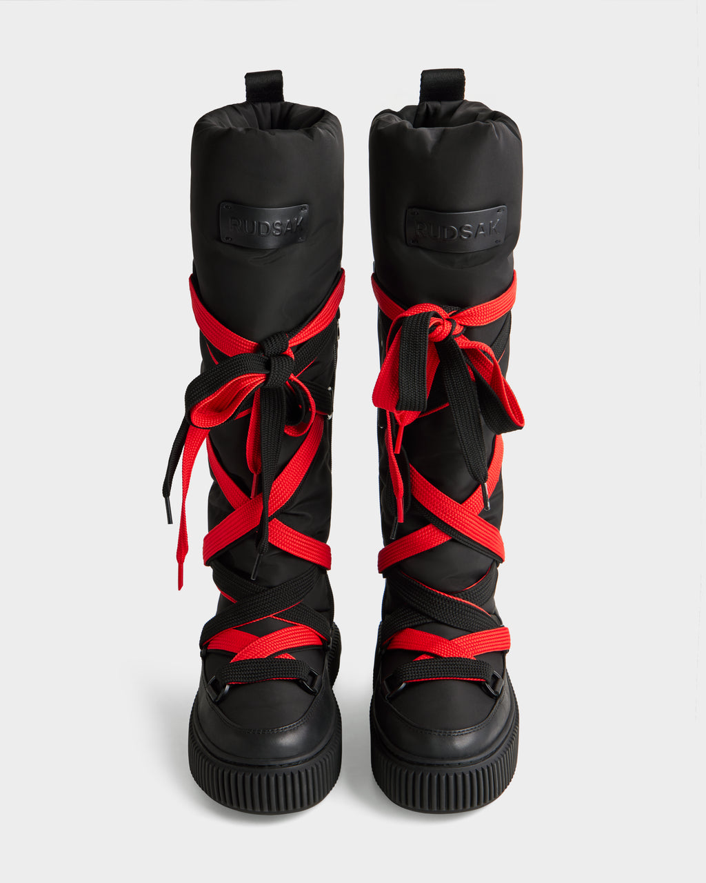 The PALERMO - 8225030 Black women’s winter boots by Rudsak feature tall insulated black design, thick red and black crisscross laces, rugged soles, and padded tops, displayed upright on a plain light background.