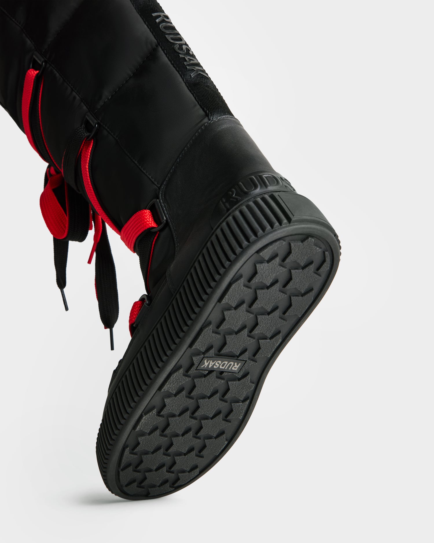 Close-up of the Rudsak PALERMO - 8225030 Black insulated women’s winter boot with red laces, featuring a textured rubber sole embossed with RUDSAK and a water-resistant tread pattern, shown slightly tilted.