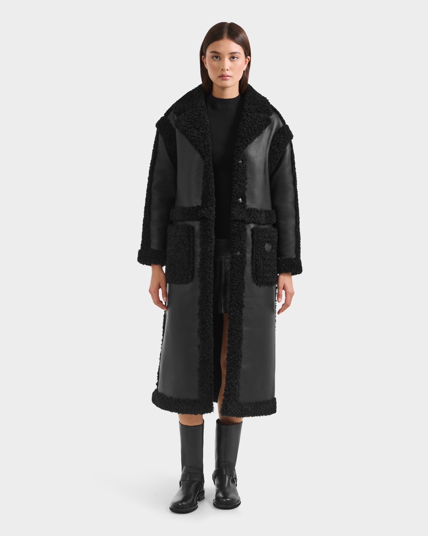 A woman stands facing forward against a plain white background, wearing the Rudsak PALOMA - 8125675 Black long faux shearling coat with large lapels and patch pockets, styled with black boots and a black outfit underneath.