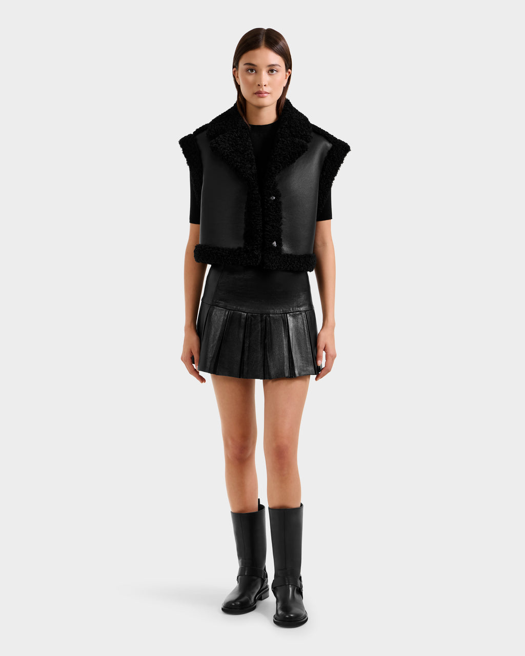 A woman faces forward against a plain light background, wearing the Rudsak PALOMA - 8125675 Black faux shearling vest, paired with a black pleated mini skirt, black short-sleeve top, and knee-high boots.