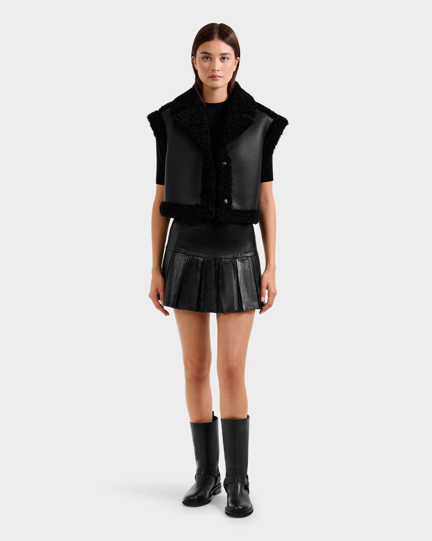 A woman faces forward against a plain light background, wearing the Rudsak PALOMA - 8125675 Black faux shearling vest, paired with a black pleated mini skirt, black short-sleeve top, and knee-high boots.