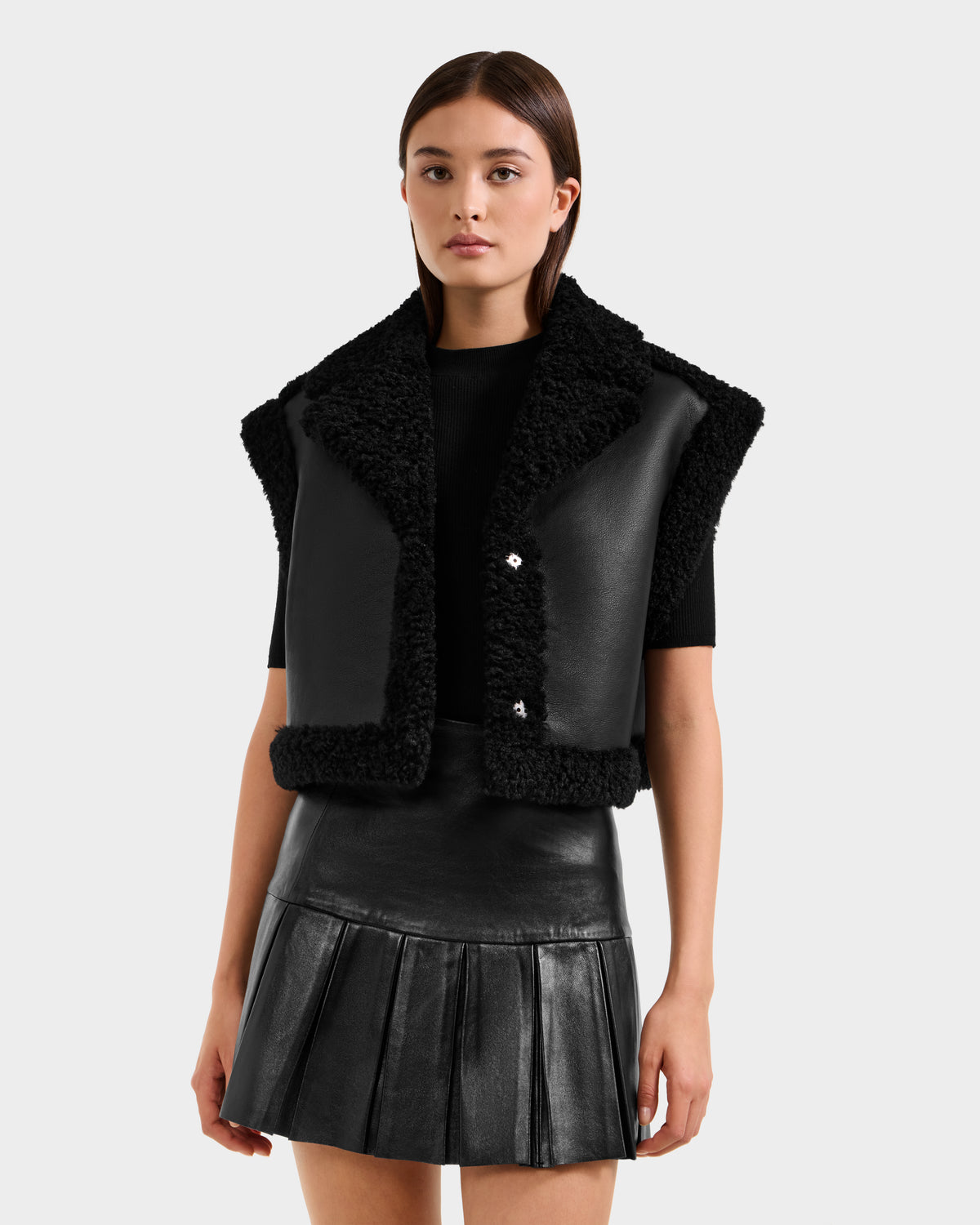 A woman wearing the Rudsak PALOMA - 8125675 Black faux shearling vest over a black top and pleated leather skirt stands against a plain light gray background.