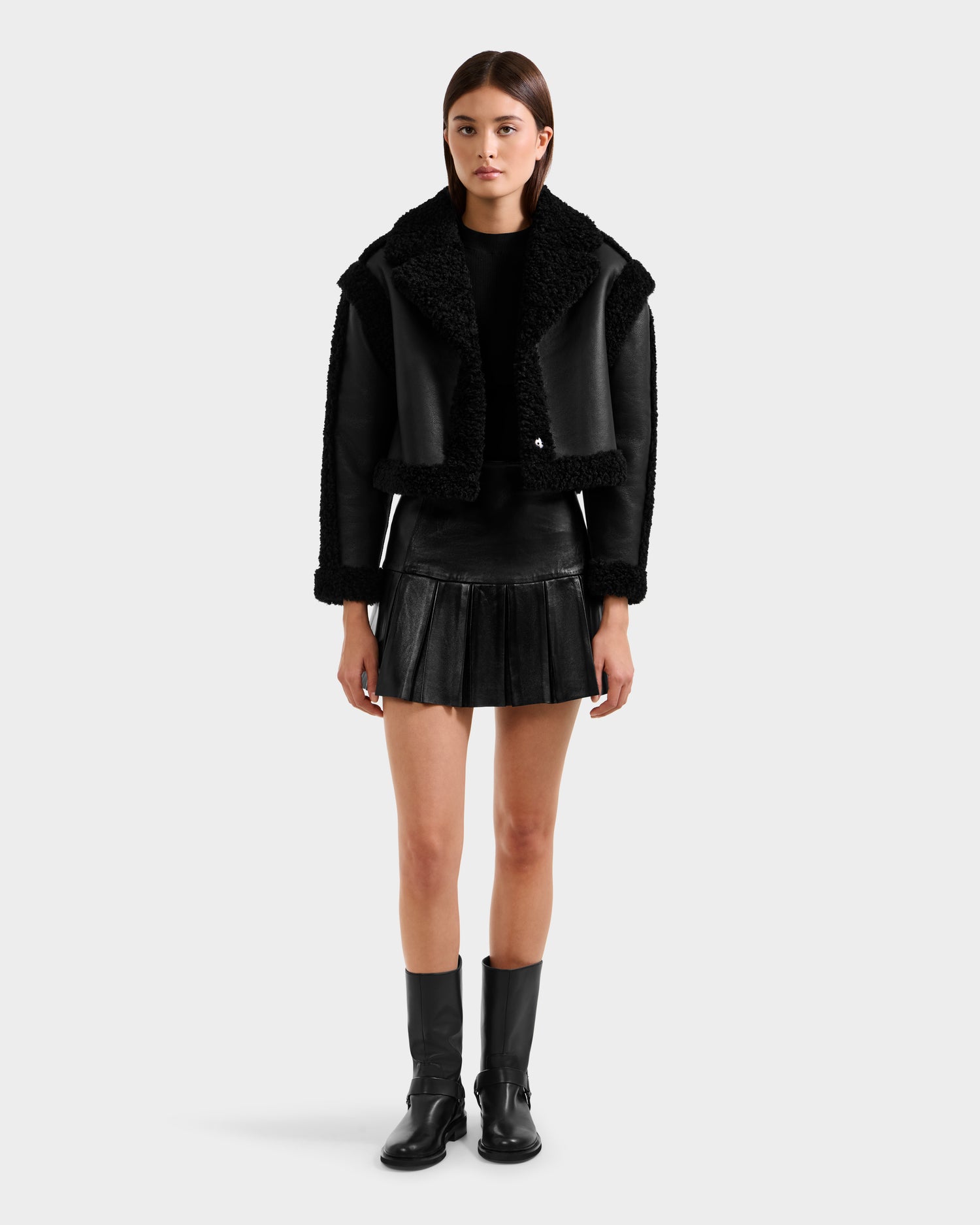 A woman stands facing forward against a plain light background, wearing the Rudsak PALOMA - 8125675 Black faux shearling jacket, paired with a black sweater, pleated mini skirt, and mid-calf boots.