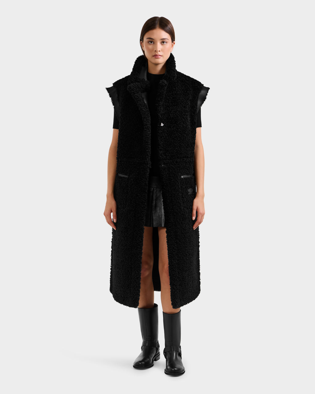 A woman faces forward, wearing the Rudsak PALOMA - 8125675 Black reversible sleeveless coat over a black outfit with knee-high boots, set against a plain light gray background.