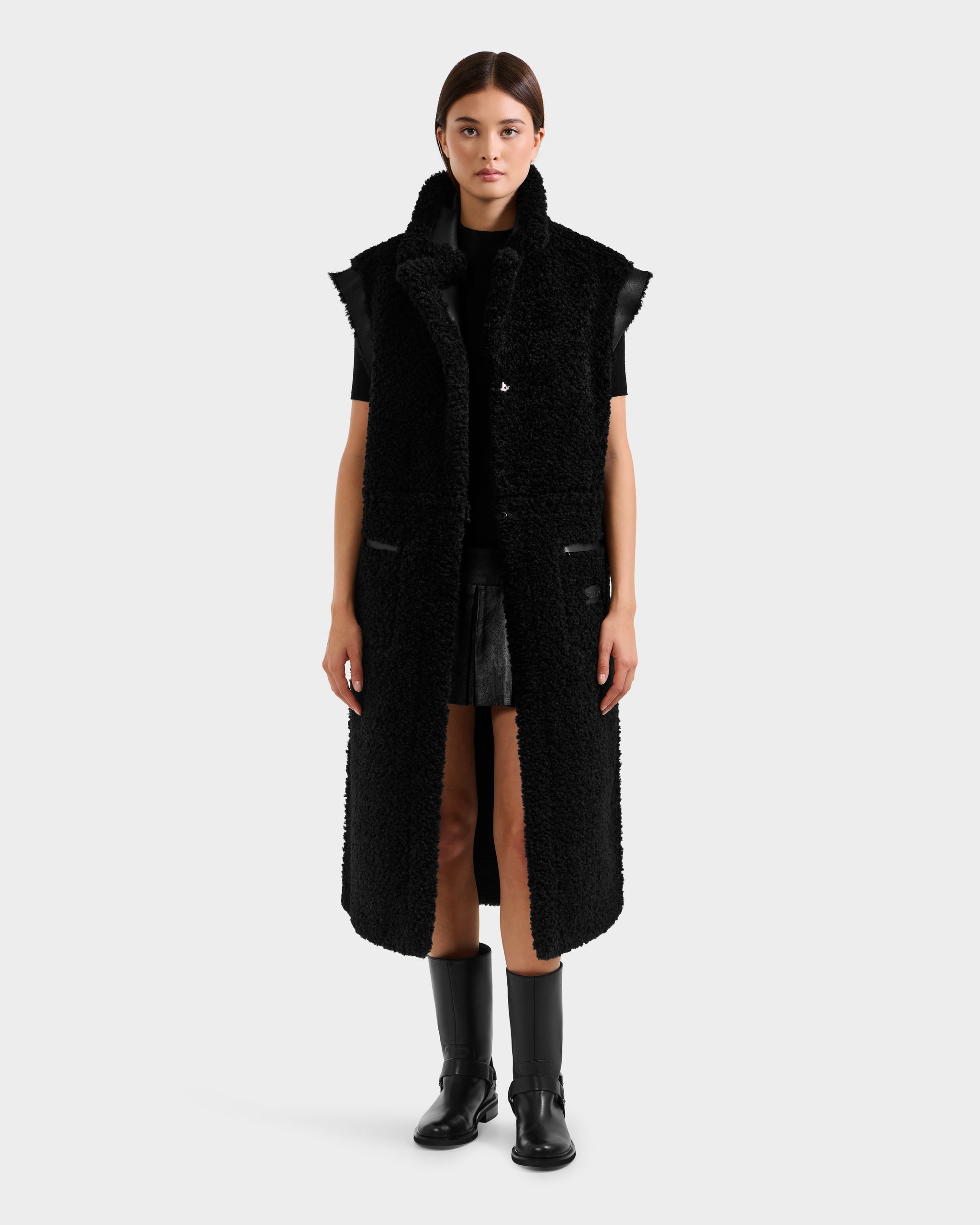A woman faces forward, wearing the Rudsak PALOMA - 8125675 Black reversible sleeveless coat over a black outfit with knee-high boots, set against a plain light gray background.