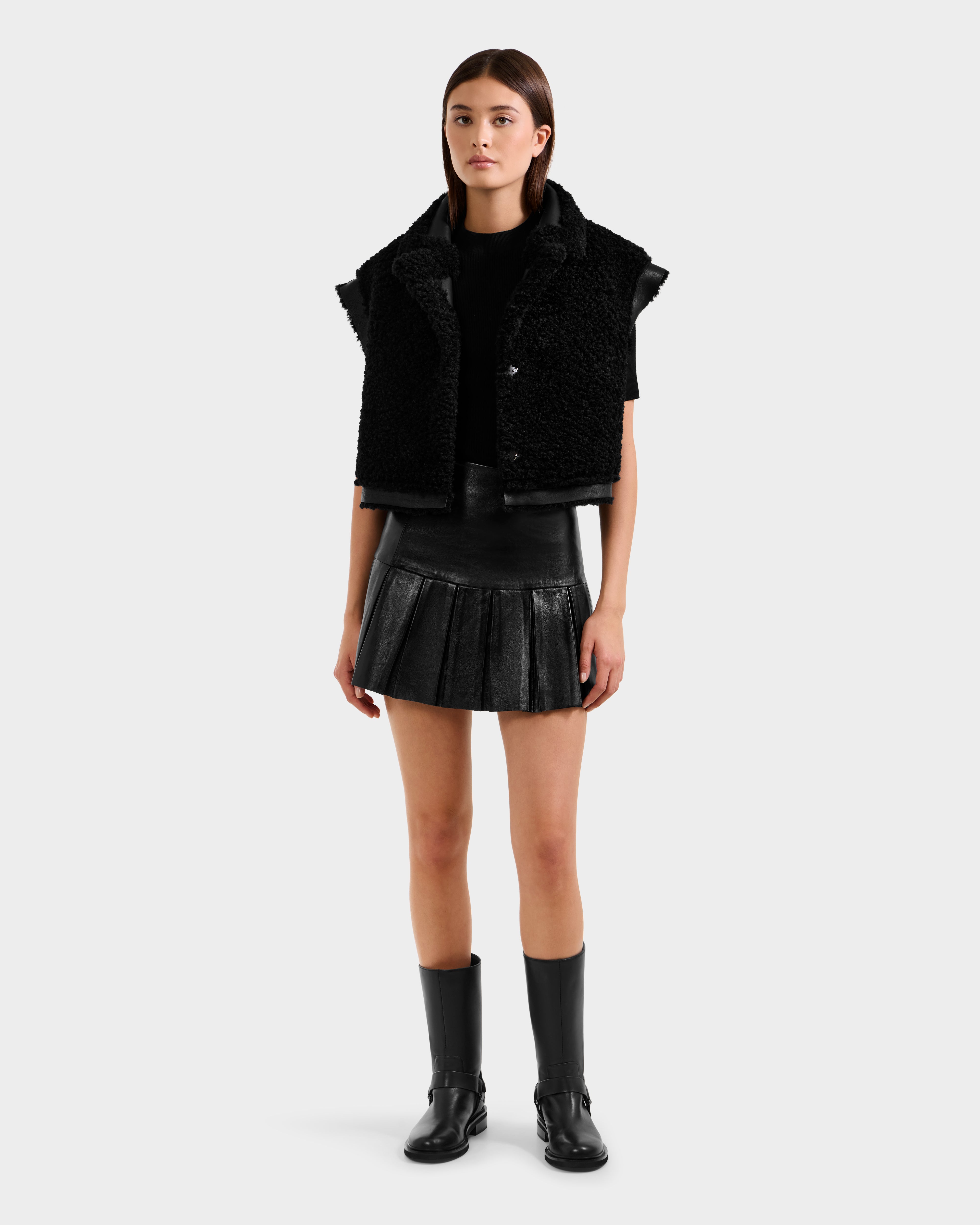 A woman with straight brown hair and a neutral expression models the Rudsak PALOMA - 8125675 Black textured faux shearling vest over a black t-shirt, pleated black leather mini skirt, and knee-high boots against a plain background.