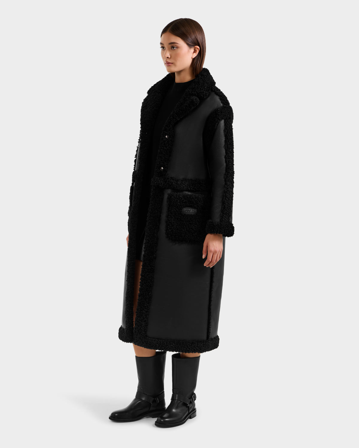 A woman with straight brown hair and a neutral expression wears the Rudsak PALOMA - 8125675 Black modular outerwear-inspired long shearling coat with textured trim and large pockets, paired with black knee-high boots against a plain light background.