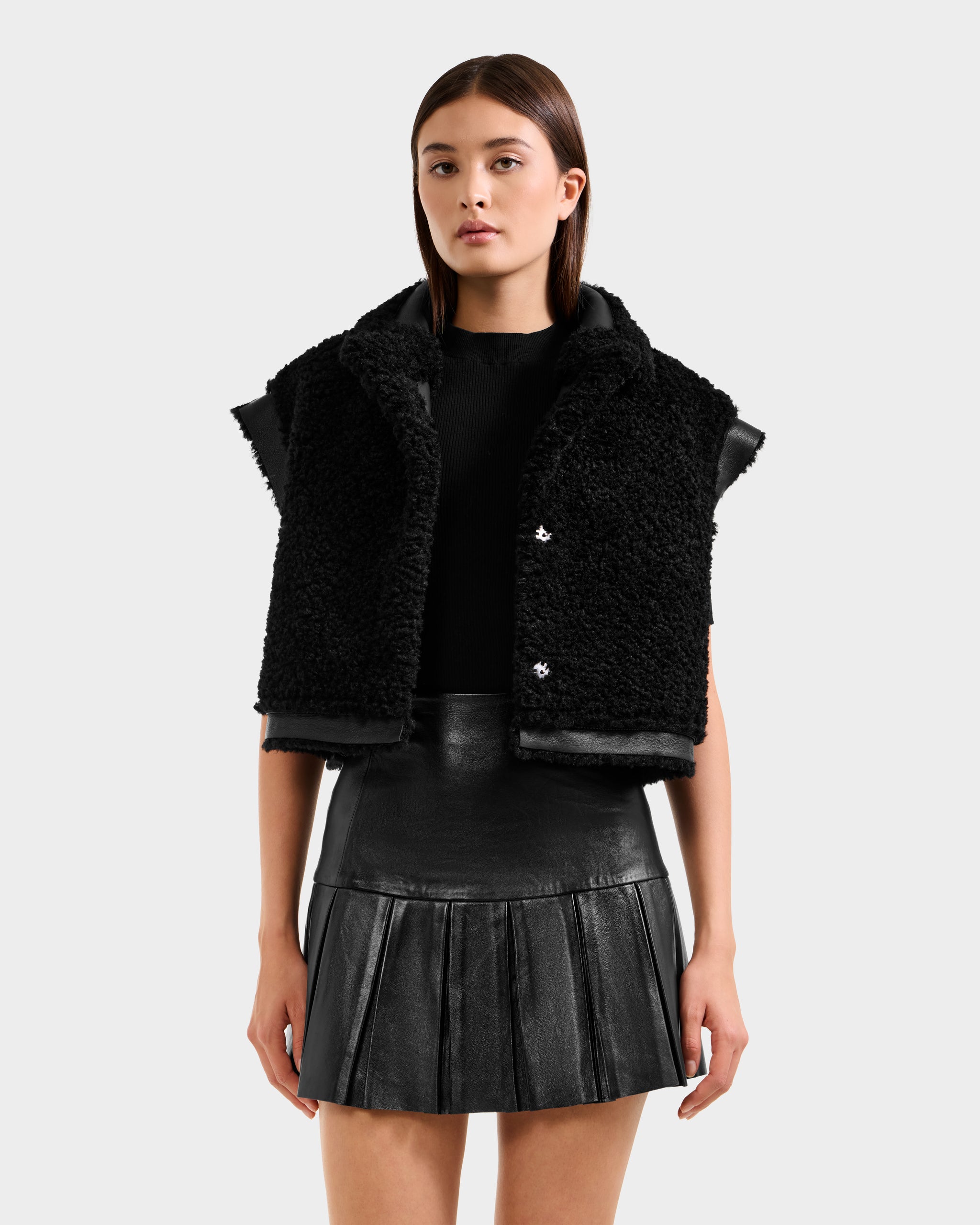 A woman with straight brown hair and a neutral expression wears the Rudsak PALOMA - 8125675 Black textured vest over a black top and pleated leather mini skirt, standing against a plain background.